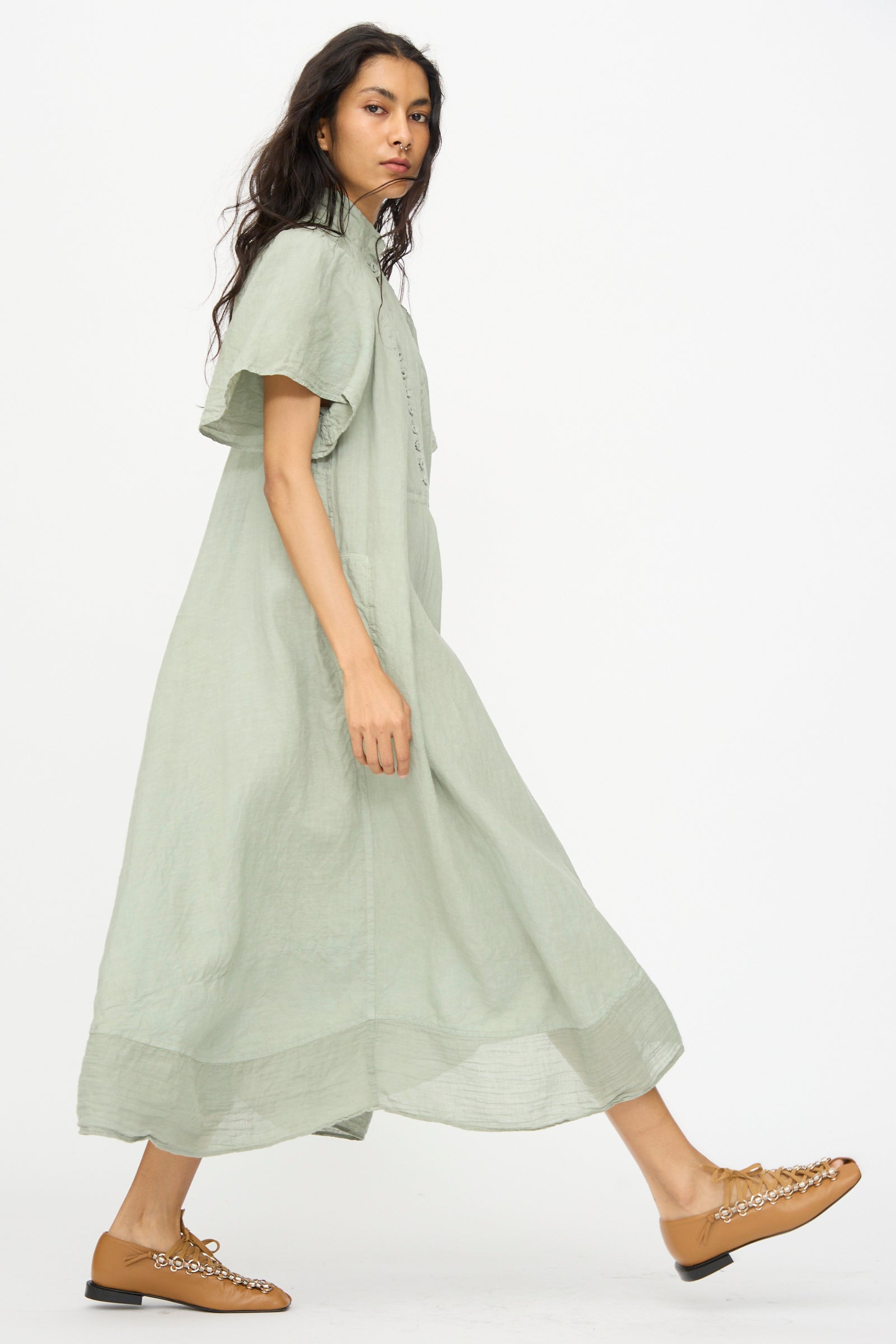 A woman wearing the Hallelujah Robe 1900's in Green Tea Dye, a light green short-sleeved linen ramie chambray maxi dress, walks sideways in tan embellished flats against a plain white background.