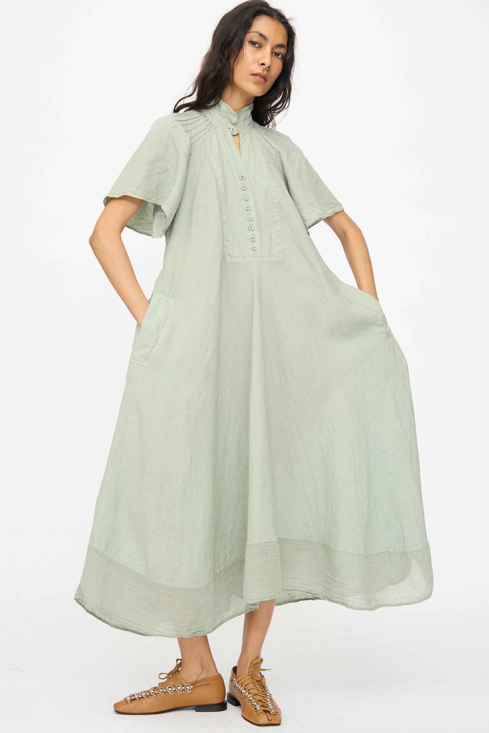 A woman wearing Hallelujah’s Robe 1900's in Green Tea Dye—a loose, short-sleeve chambray linen dress with front buttons and side pockets—pairs it with brown lace-up shoes against a plain white background.