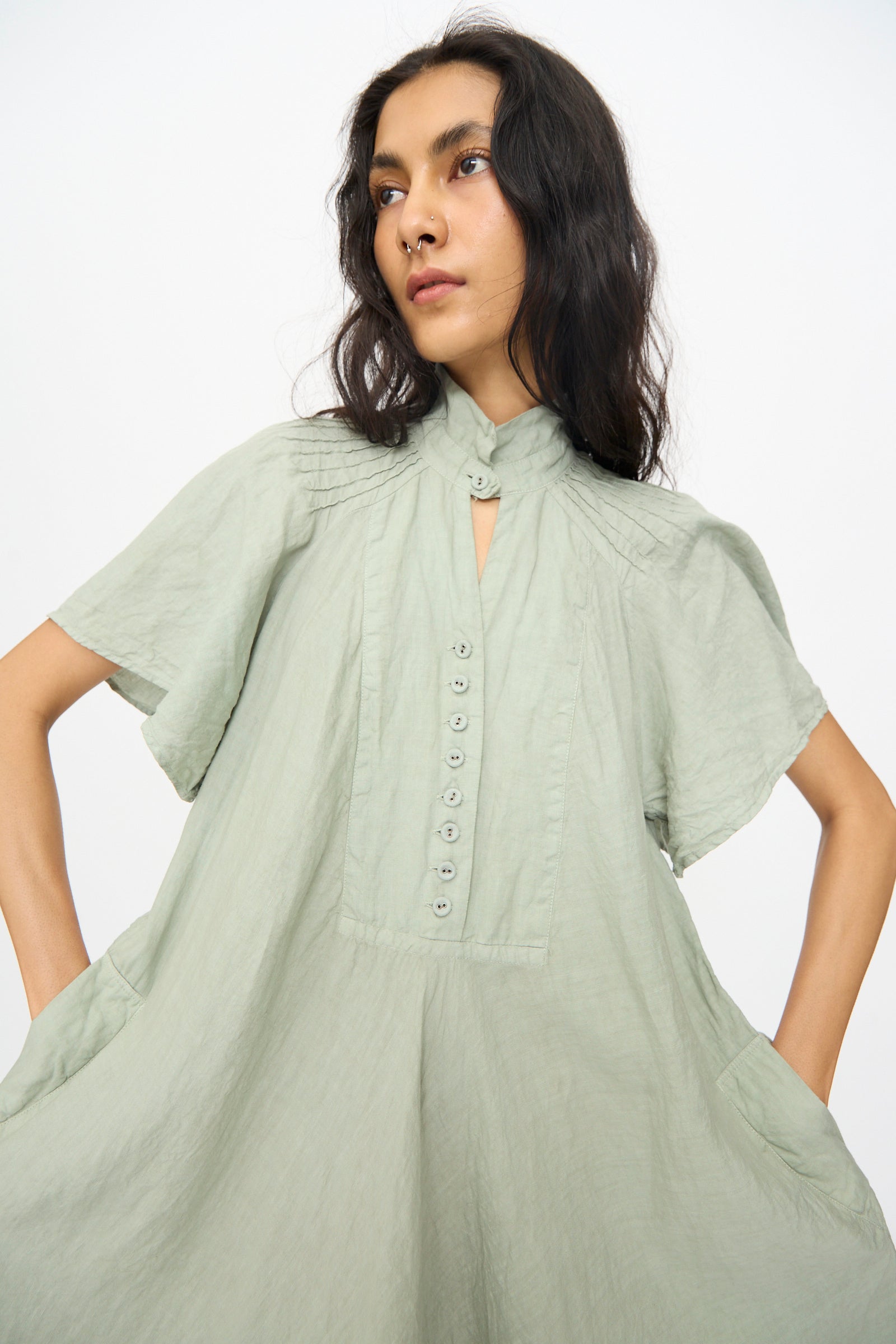 A person wears Hallelujah's Robe 1900's in Green Tea Dye, a light green, short-sleeved linen ramie chambray maxi dress with a high collar, buttoned front, and side pockets, standing against a plain white background.