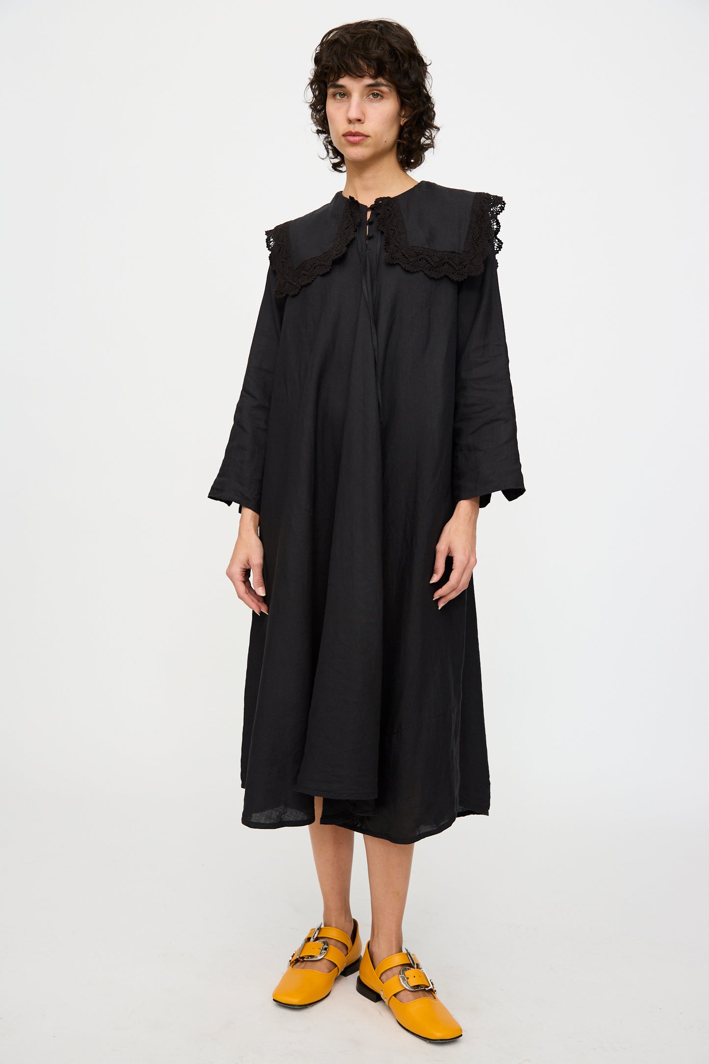 Wearing Hallelujah's Robe Religieuse col carré in black, featuring an A-line silhouette and large lace collar, a person pairs the ensemble with yellow shoes against a plain white background.