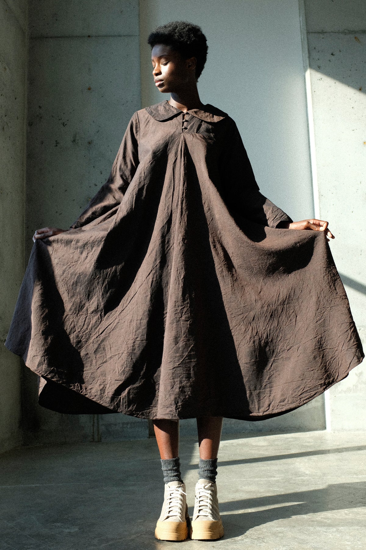 A woman holding up the sides of the Hallelujah Linen Wool Robe à Col Claudine in Brown standing in a sunlight studio.