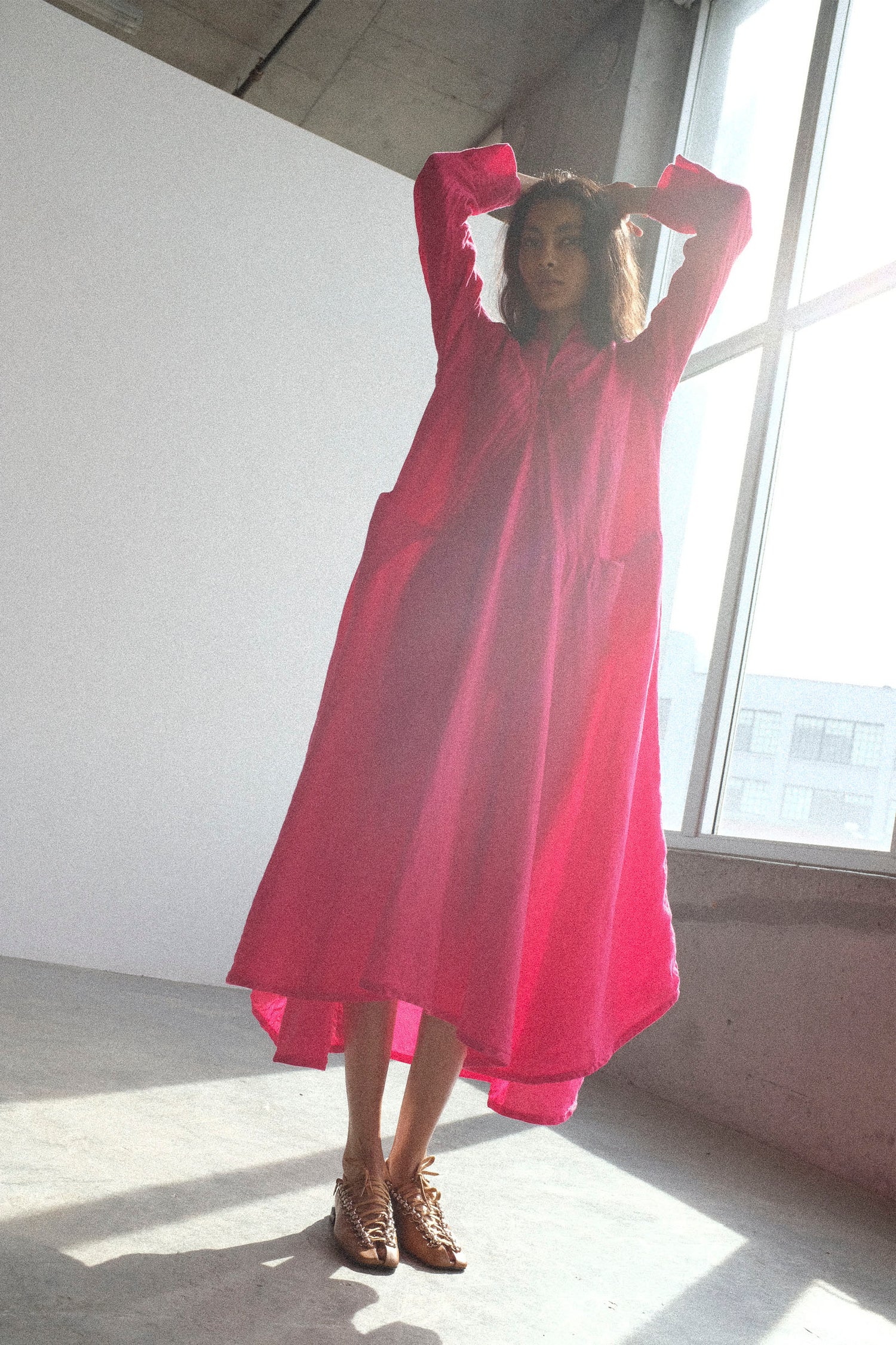 A woman wearing Hallelujah's Cotton Linen Robe de Femme de Chambre in Cherry Red, standing in a room with concrete walls and large windows.