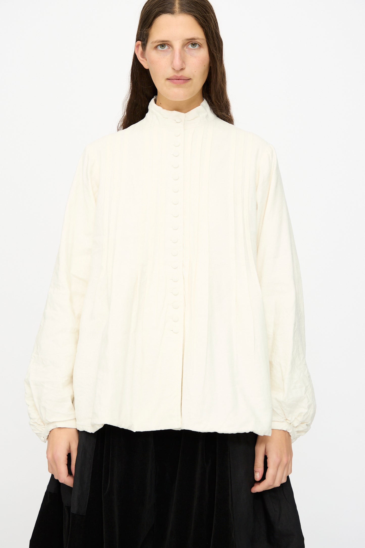 A person with long brown hair wears the Hallelujah Silk and Linen Chemise de Baptême in White, a loose-fitting blouse with a high collar and covered buttons, paired with a black skirt.