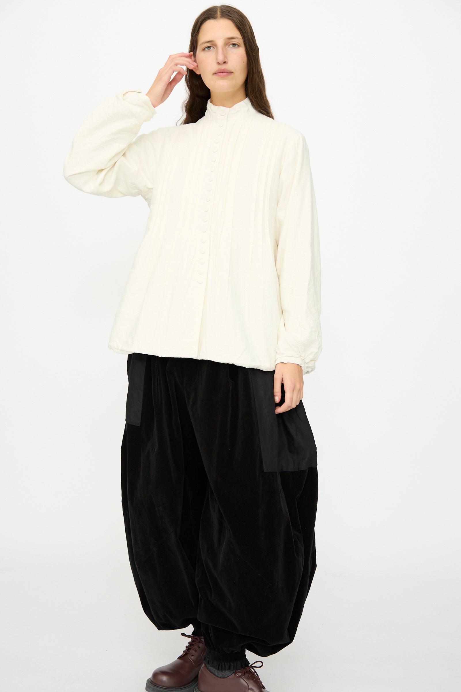 A person stands against a plain background wearing the Hallelujah Silk and Linen Chemise de Baptême in White, paired with wide black pants and dark shoes.