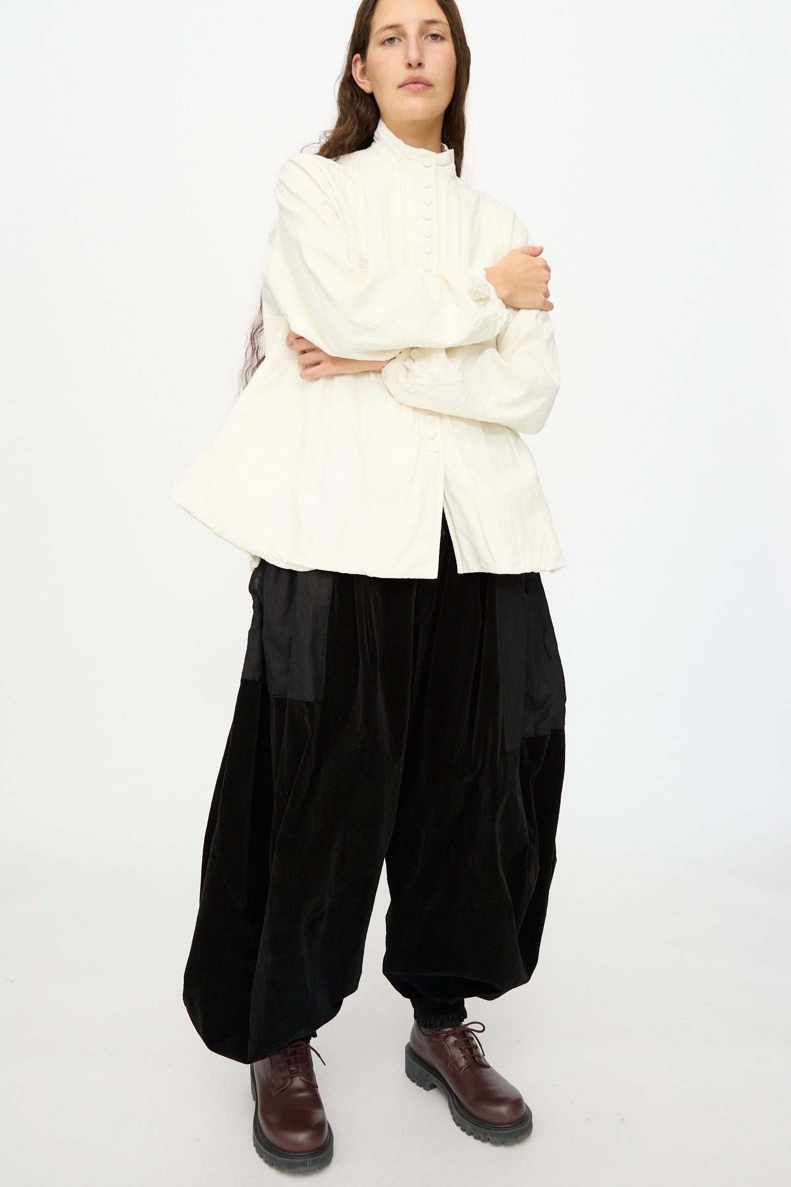 A person stands with arms crossed in a plain white space, wearing the Hallelujah Silk and Linen Chemise de Baptême in White, loose black pants, and brown lace-up boots.