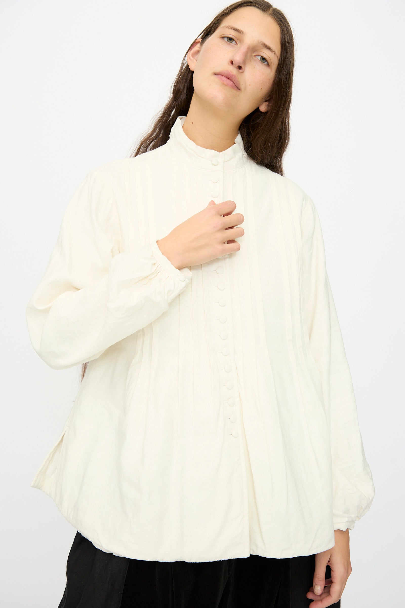 A person with long brown hair models the Hallelujah Silk and Linen Chemise de Baptême in White, a loose-fitting blouse with a high collar and front buttons, standing against a plain white background.