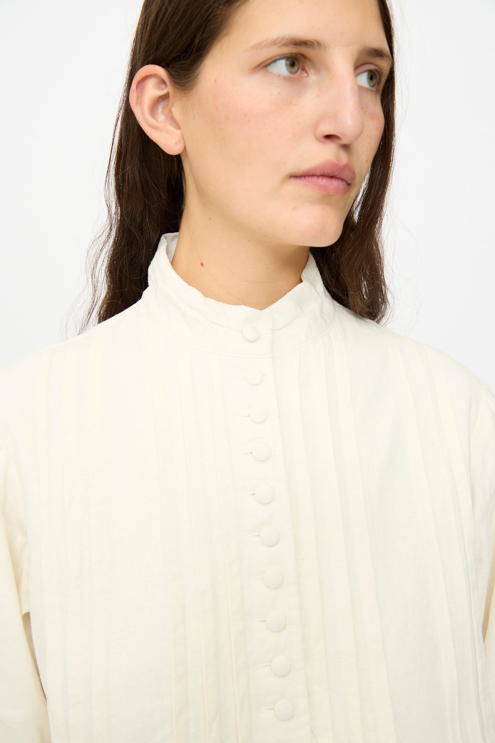 A person with long brown hair wears the Hallelujah Silk and Linen Chemise de Baptême in White, high-collared and buttoned, facing slightly right against a plain background.