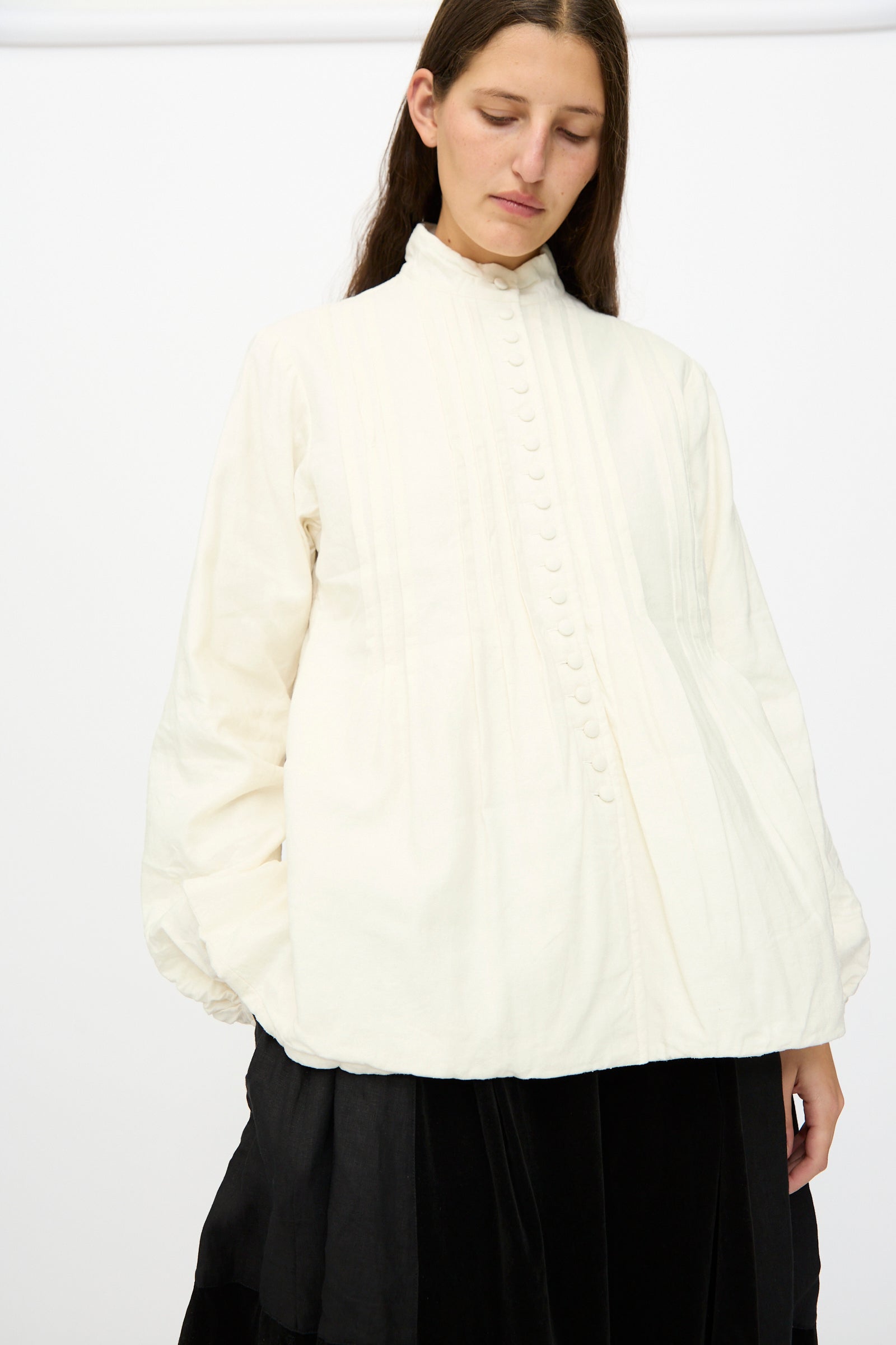 A person wears the Hallelujah Silk and Linen Chemise de Baptême in White—a high-neck, long-sleeved blouse with pleats and buttons—paired with a black skirt, standing against a plain white background.