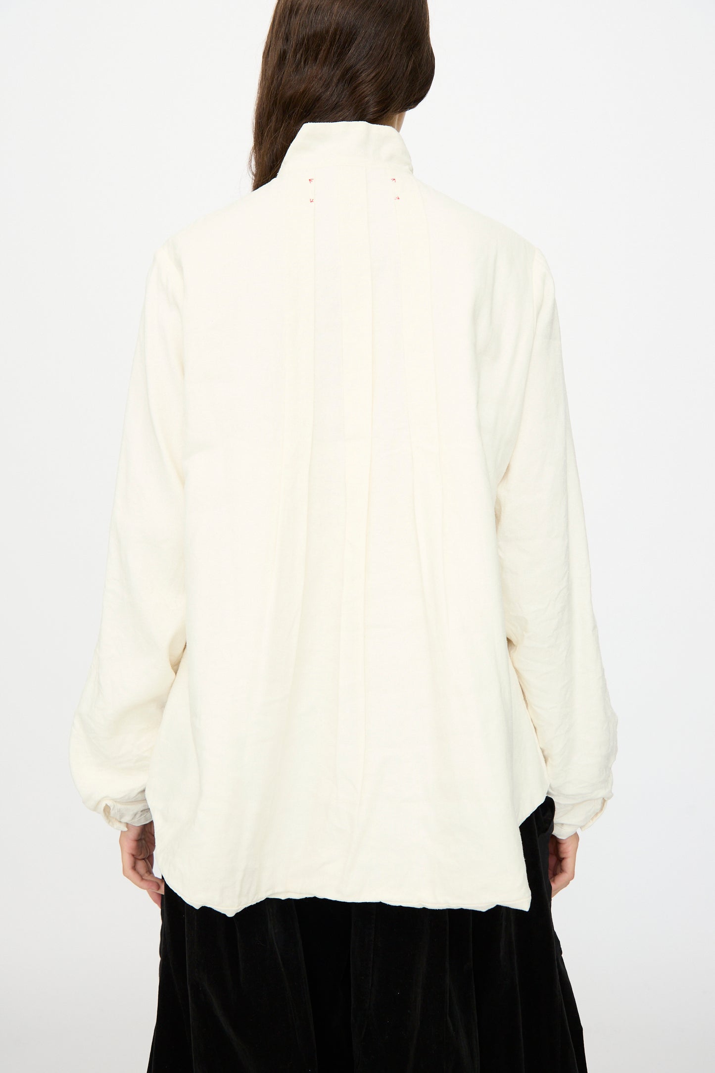 A person is seen from behind wearing the Hallelujah Silk and Linen Chemise de Baptême in White, a loose, long-sleeved blouse with a high collar, crafted in Japan and photographed against a plain background.