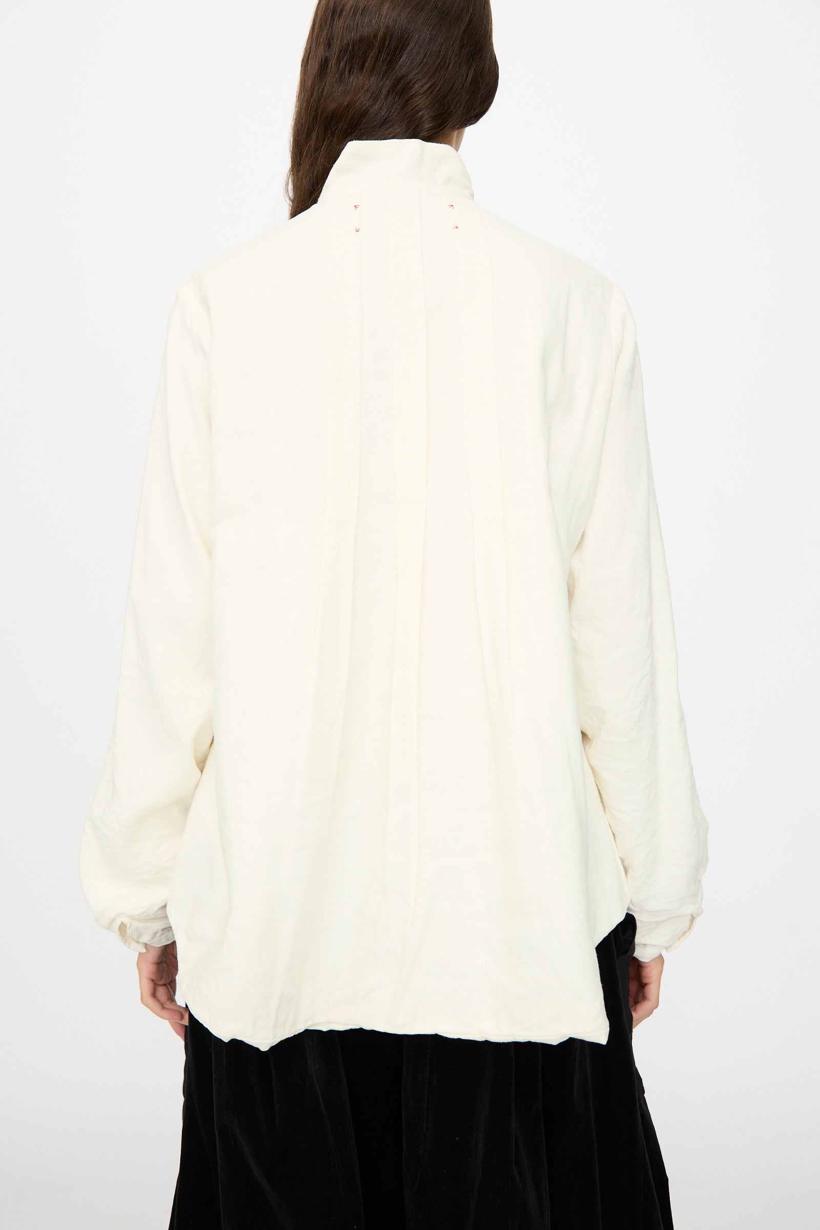 A person is seen from behind wearing the Hallelujah Silk and Linen Chemise de Baptême in White, a loose, long-sleeved blouse with a high collar, crafted in Japan and photographed against a plain background.