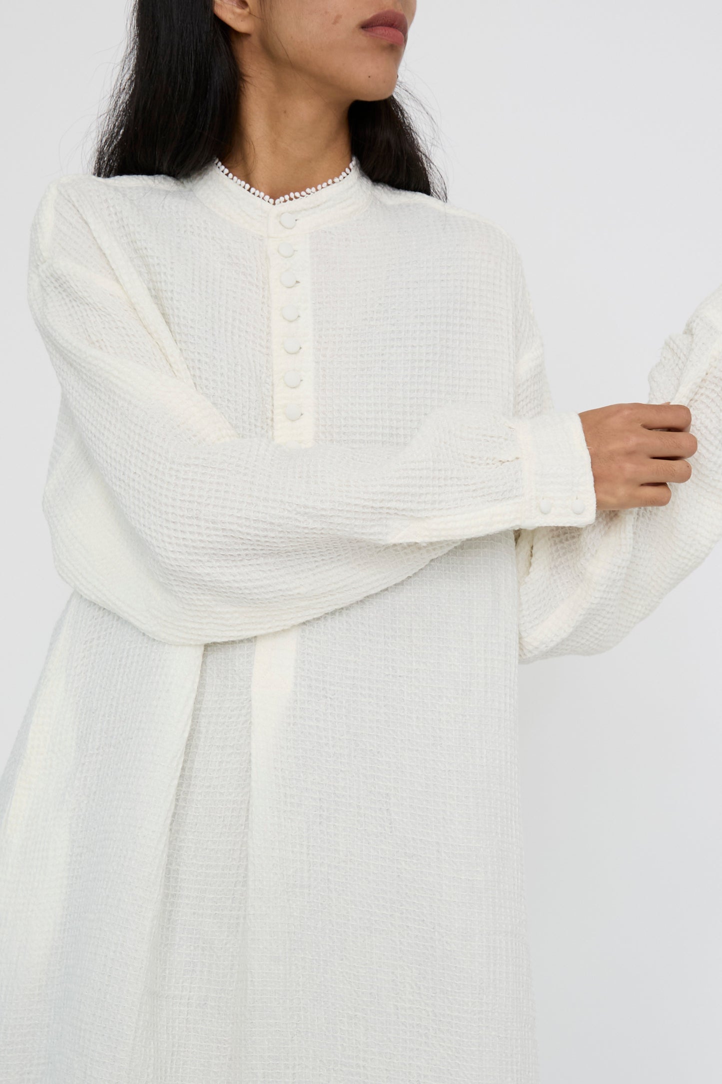 A person adjusts their sleeve wearing the Waffle Linen Robe Chemise in Milky White by Hallelujah, which features long sleeves, button-up design, and a relaxed fit in textured linen against a plain background.