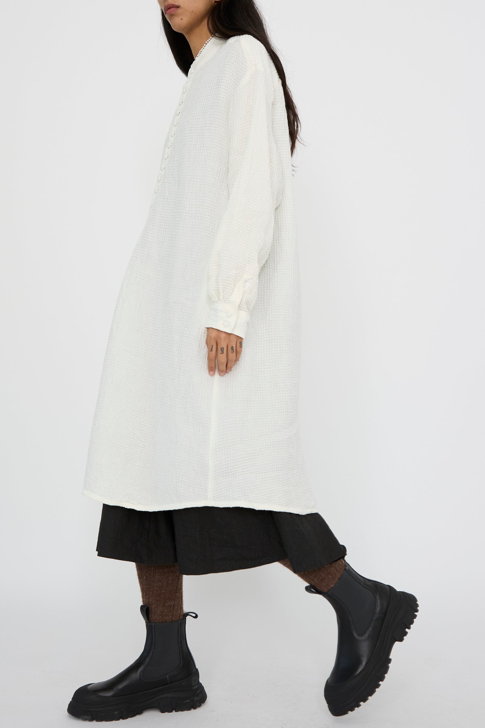 Dressed in Hallelujah's Waffle Linen Robe Chemise in Milky White, paired with a black dress and boots, an individual stands against a plain background, facing left.