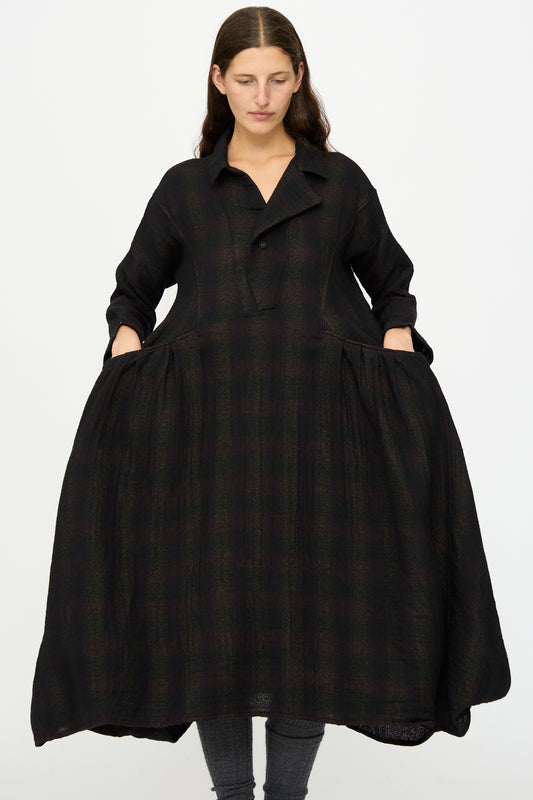 A woman stands against a white background wearing the Hallelujah Wool Cotton Robe de Berger in Black Mix, a loose-fitting, long-sleeve dress made in Japan with dark plaid, pockets, and paired with grey leggings.