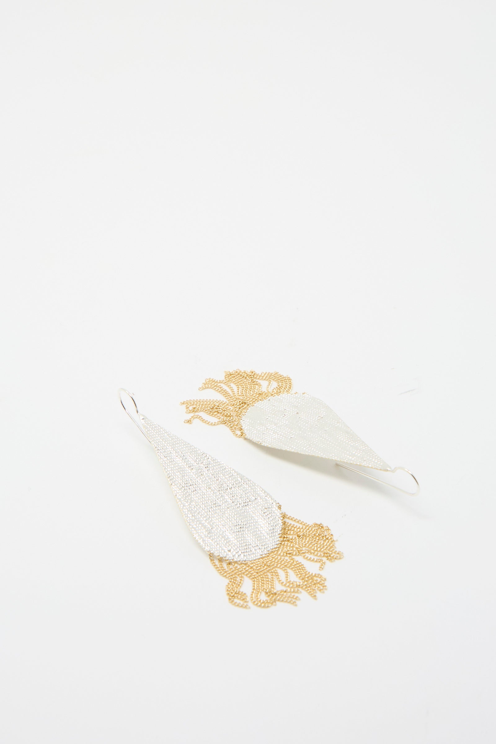 The Drip Earrings by Hannah Keefe are a pair of silver teardrop earrings, adorned with a brass chain fringe and fringed ends, presented on a white background. 