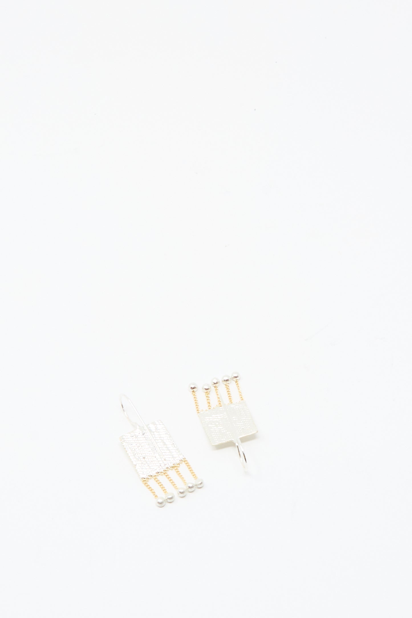 The Bauble Earrings in Brass and Silver by Hannah Keefe, featuring two rectangular handmade pieces with metallic chains and small ball ends, are displayed on a plain white background.