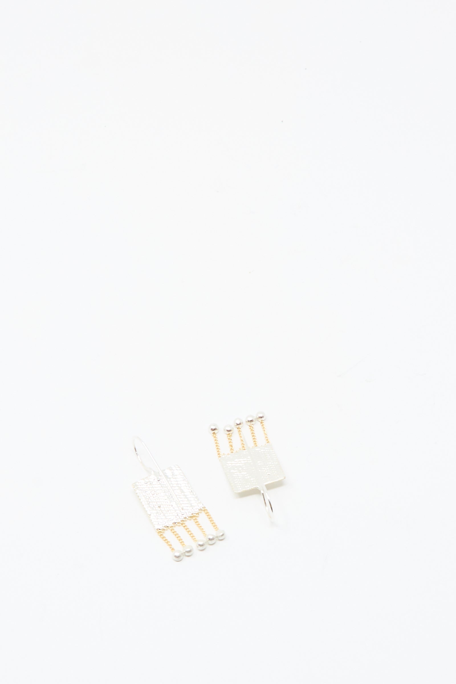 The Bauble Earrings in Brass and Silver by Hannah Keefe, featuring two rectangular handmade pieces with metallic chains and small ball ends, are displayed on a plain white background.