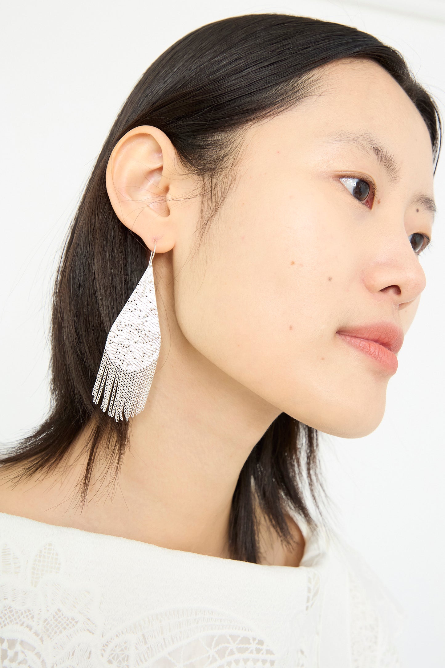 A person with shoulder-length dark hair wears a white, lace-detailed garment and a pair of Hannah Keefe's Drip Earrings in Silver, featuring large teardrops with silver chain fringe, as they gaze slightly away.