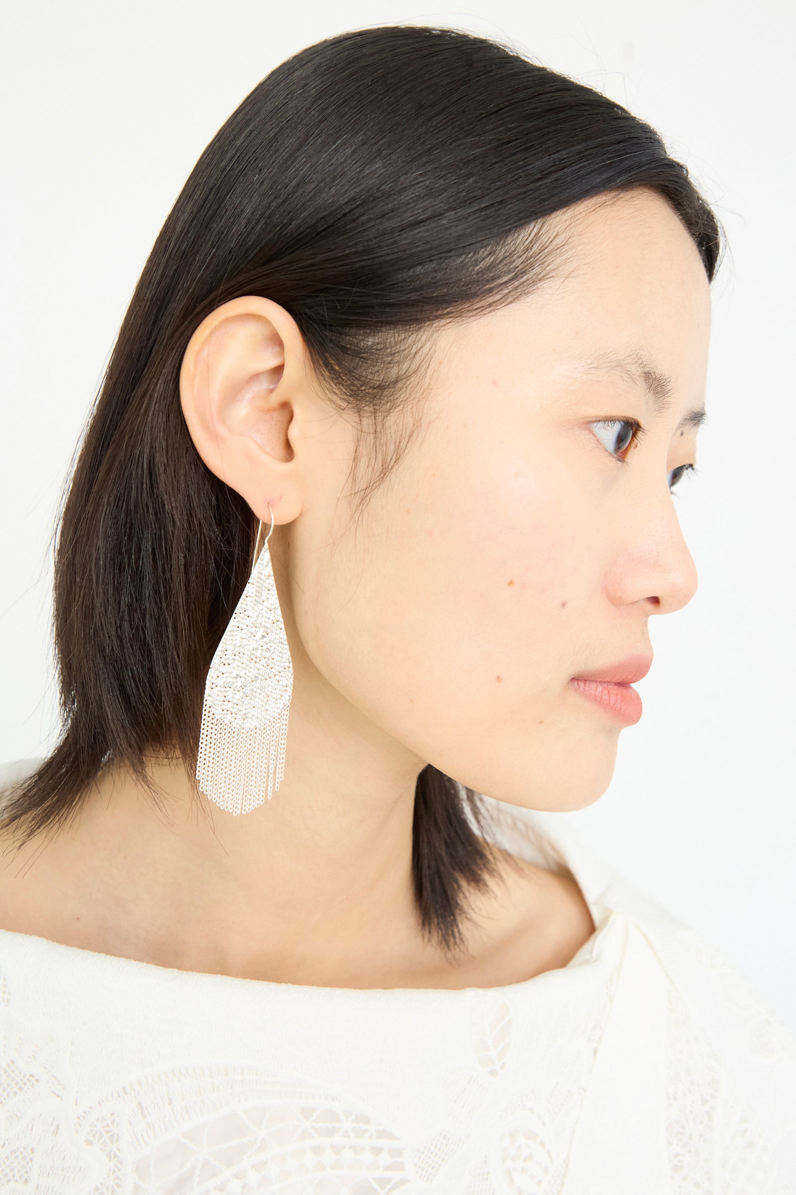 Against a white background, a person with short dark hair is viewed in profile wearing intricate Hannah Keefe Drip Earrings in Silver featuring a handmade silver chain fringe.