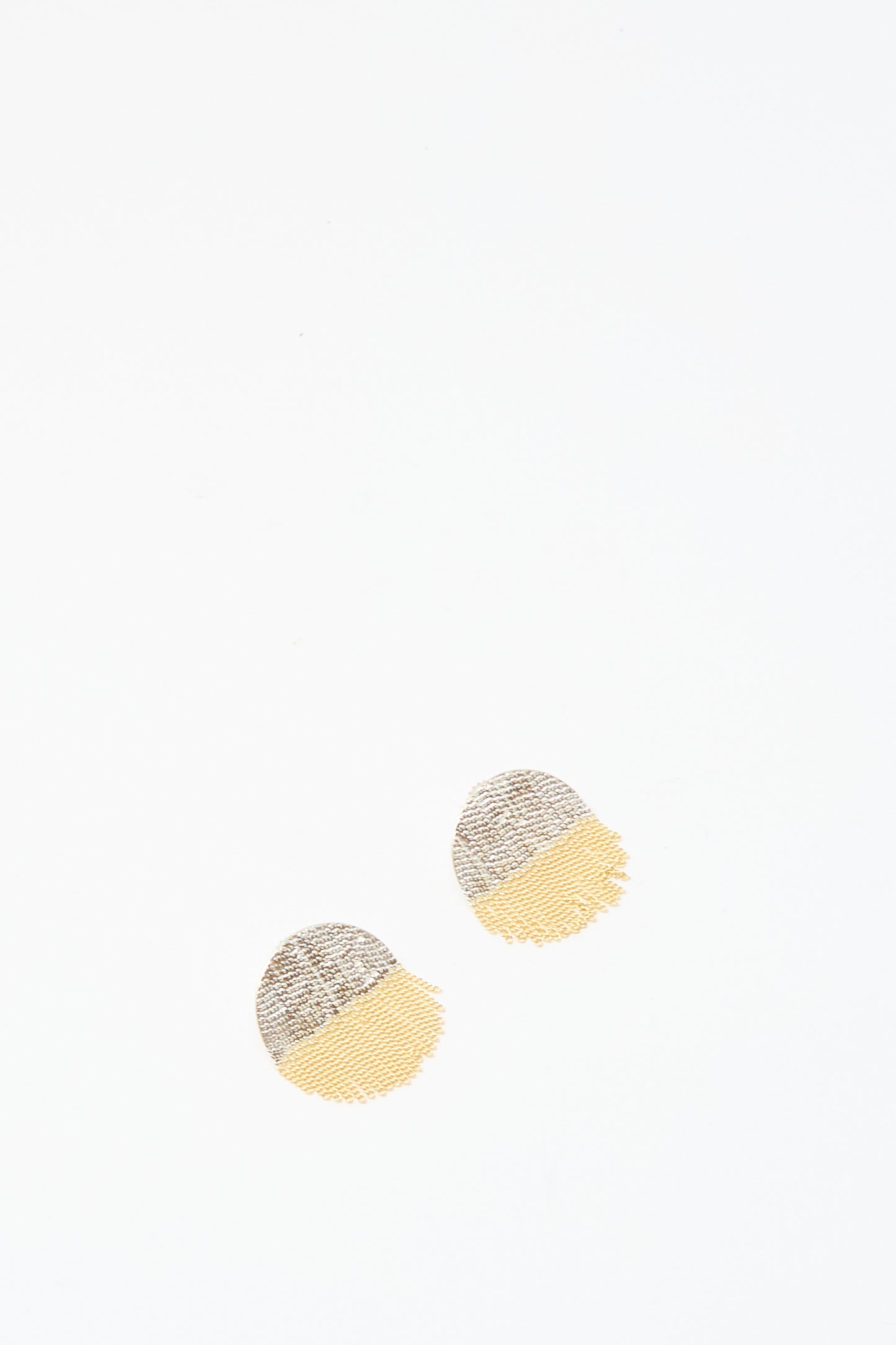 Two Half Circle Studs in Brass and Silver by Hannah Keefe, featuring textured round shapes with gray silver tops and yellow brass bottoms, are placed near the center on a white background.
