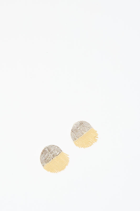Two Half Circle Studs in Brass and Silver by Hannah Keefe, featuring textured round shapes with gray silver tops and yellow brass bottoms, are placed near the center on a white background.