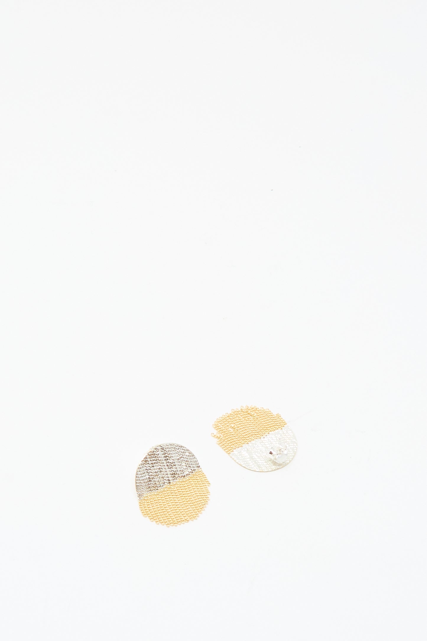 The Half Circle Studs in Brass and Silver by Hannah Keefe are paired with two small, circular beige and gray fabric patches with stitched patterns, arranged on a white background for a charming display.