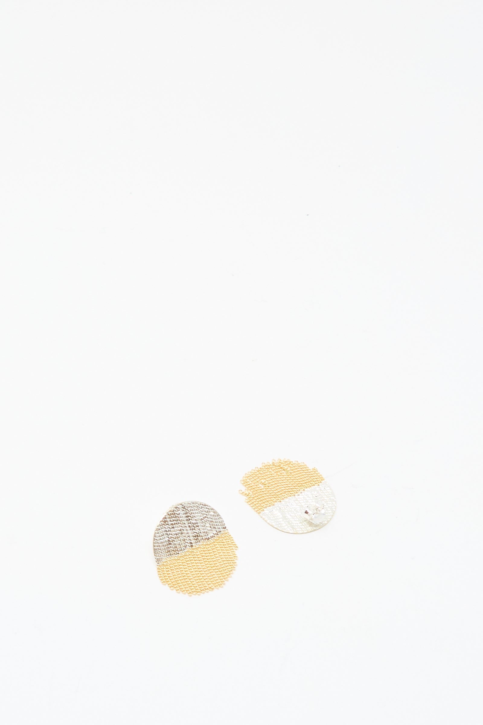 The Half Circle Studs in Brass and Silver by Hannah Keefe are paired with two small, circular beige and gray fabric patches with stitched patterns, arranged on a white background for a charming display.