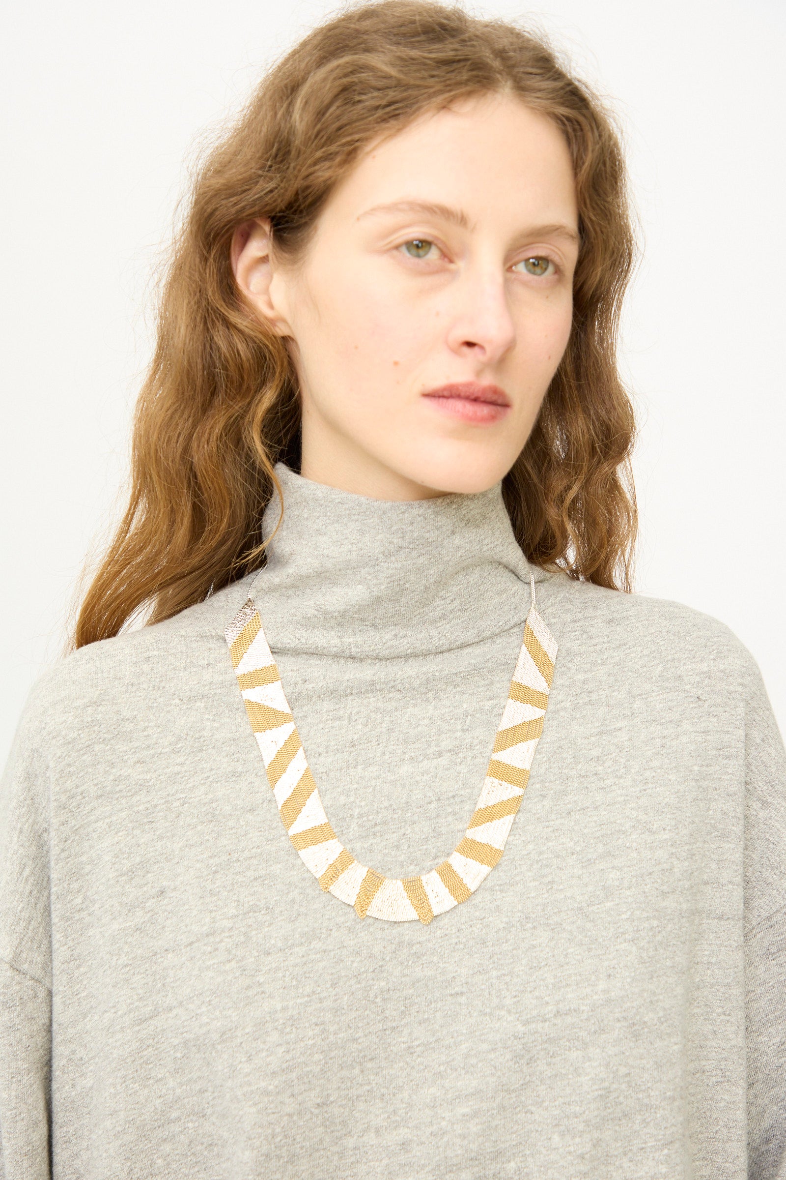 A person with long wavy hair wears a light grey turtleneck and the Hannah Keefe Sunny Necklace in Brass and Silver, featuring geometric silver triangles, standing against a plain background.