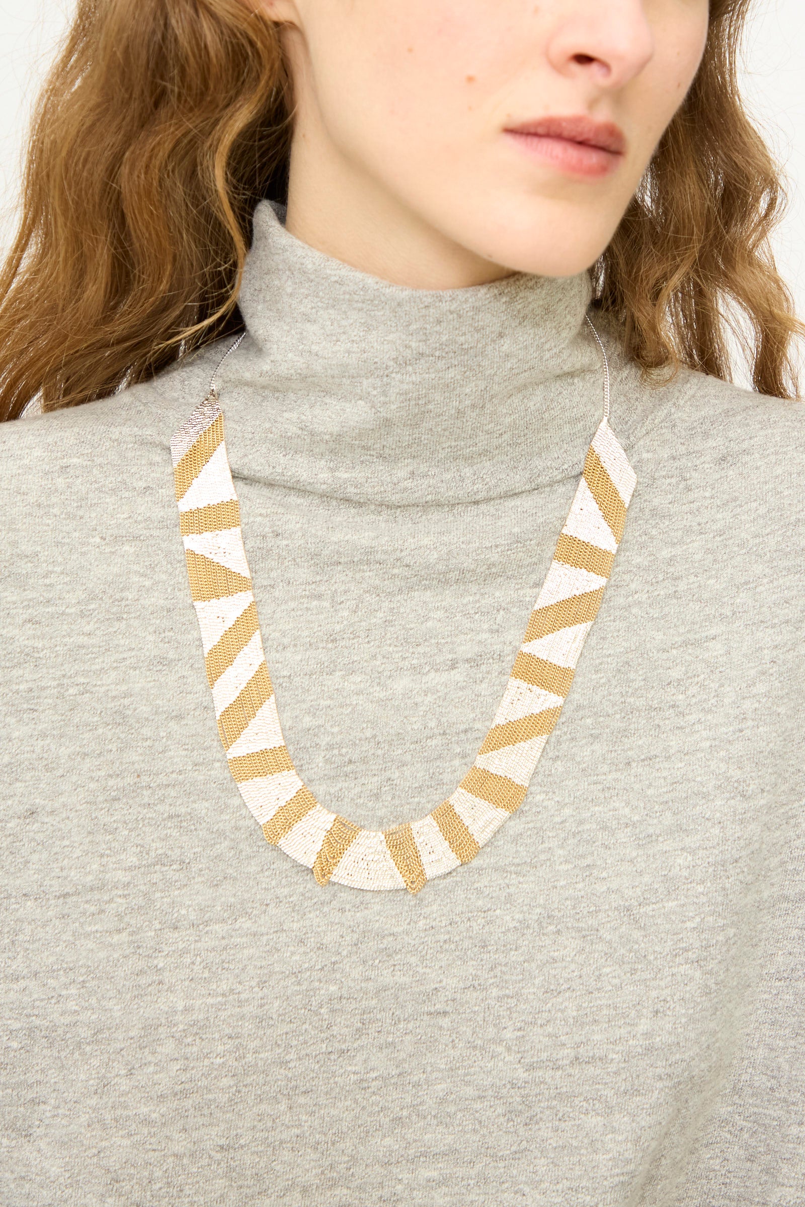 A person wearing a light gray turtleneck sweater and the Hannah Keefe Sunny Necklace in Brass and Silver, featuring handmade alternating gold and white triangular segments on a brass chain.