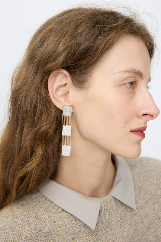 A woman with light brown hair wears Hannah Keefe's Train Earrings in Brass and Silver—rectangular, textured, and handmade—paired with a beige collared sweater, facing right against a plain background.