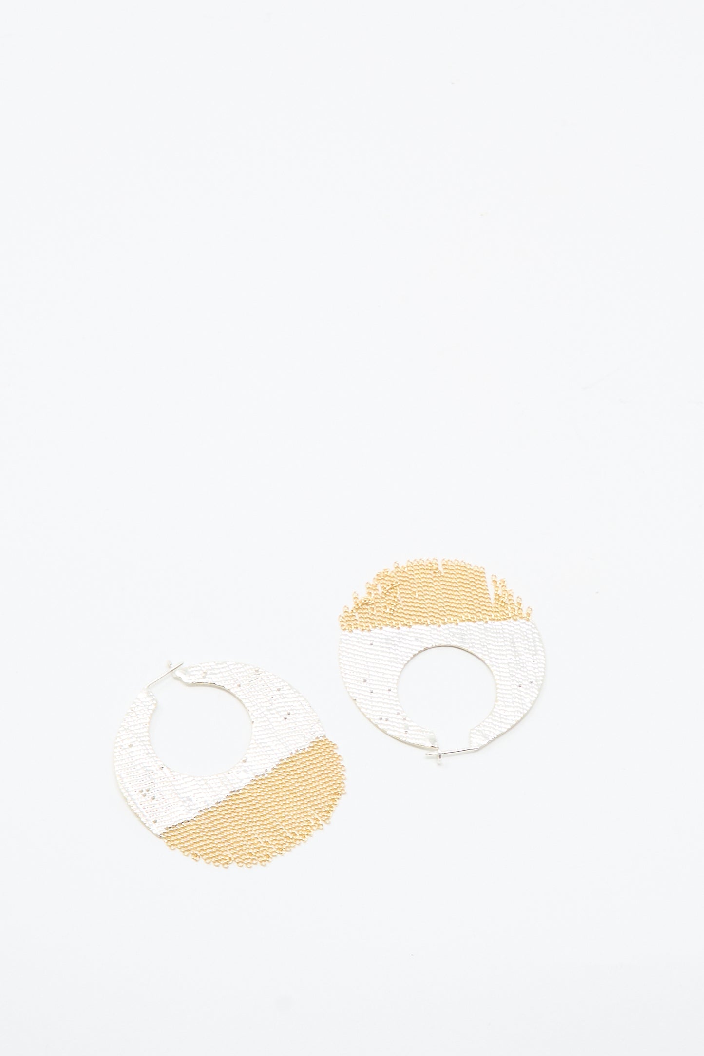 The True Hoops in Brass and Silver by Hannah Keefe are circular earrings featuring white and gold beaded sections, displayed on a plain white background.