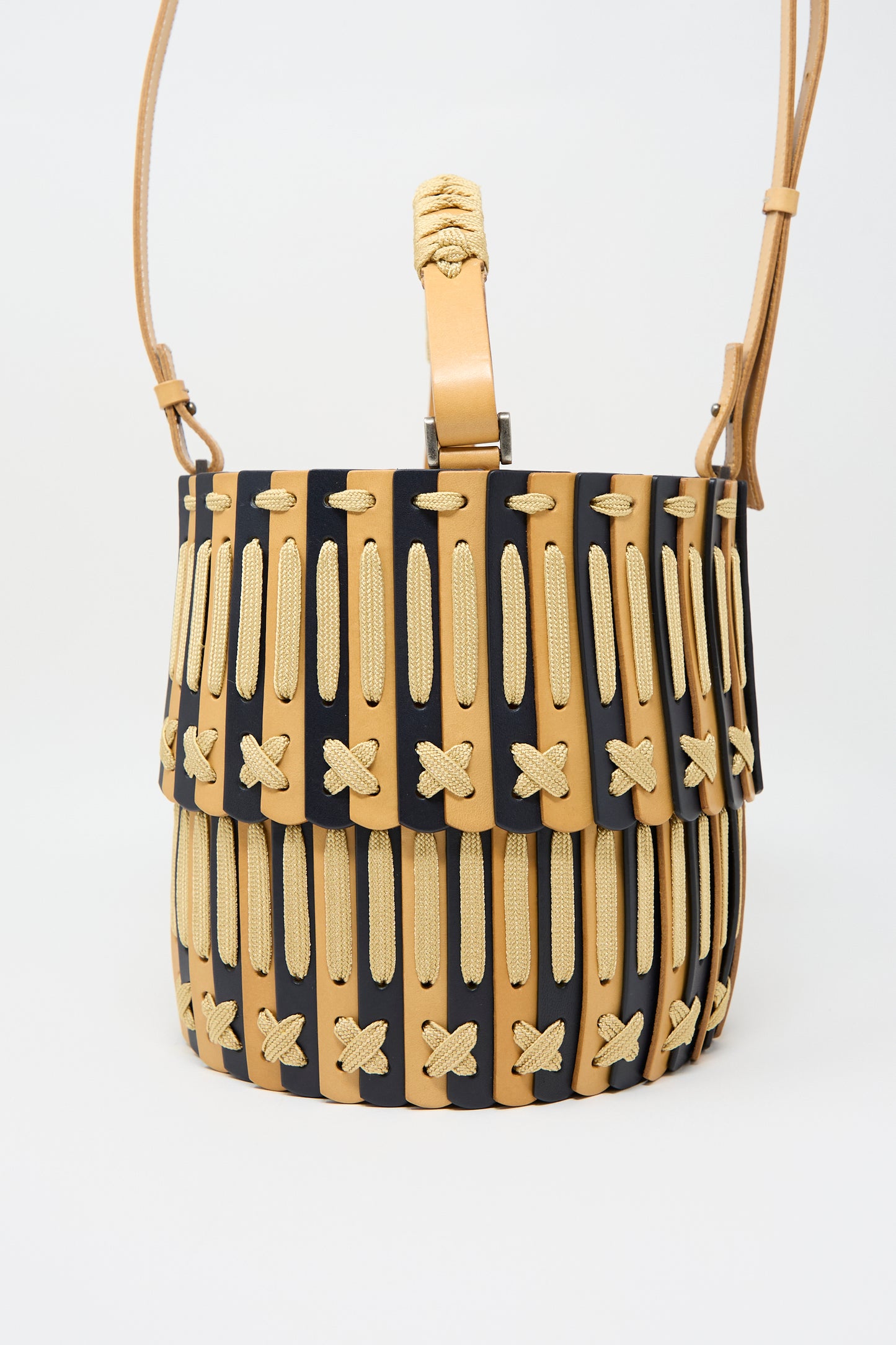 A Leather Bucket Bag in Natural and Midnight Blue by Hatori, featuring a cylindrical shape and a woven beige and black design with a flap closure, complemented by an adjustable strap made from vegetable tanned leather.