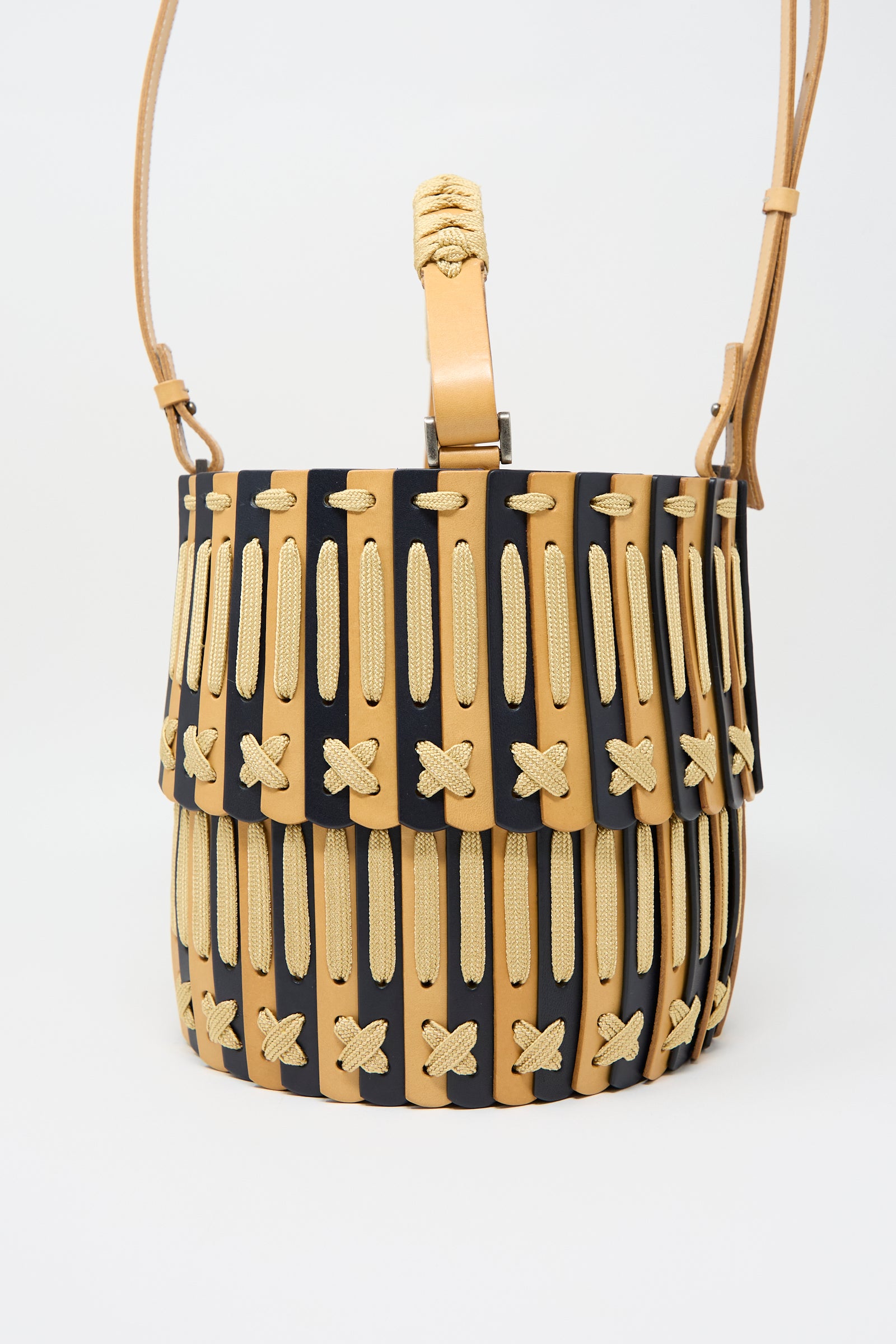 A Leather Bucket Bag in Natural and Midnight Blue by Hatori, featuring a cylindrical shape and a woven beige and black design with a flap closure, complemented by an adjustable strap made from vegetable tanned leather.