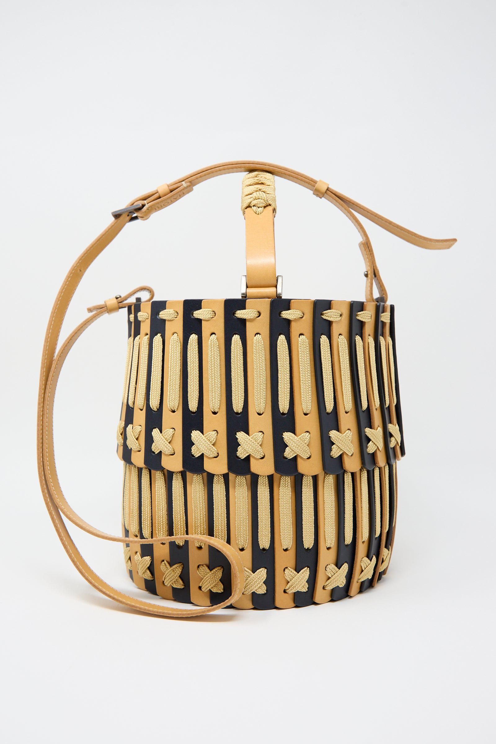 The "Leather Bucket Bag in Natural and Midnight Blue" by Hatori is a handcrafted leather bucket bag with a tan and black woven design, featuring a single handle and an adjustable shoulder strap made from vegetable-tanned leather.