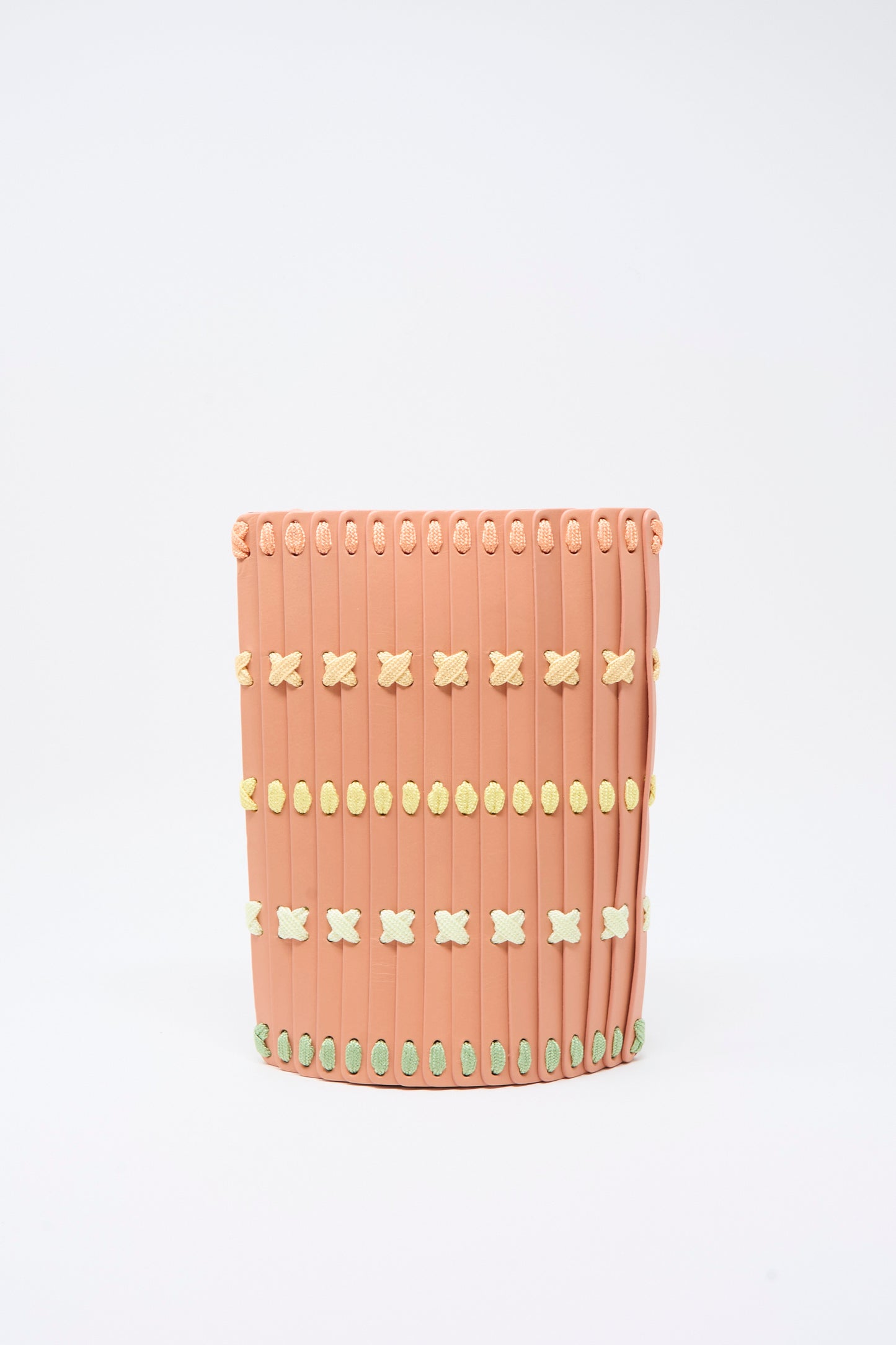 A cylindrical, pink leather crossbody bag by Hatori, featuring vertical ridges and horizontal rows of small yellow, green, and white X-shaped multi lacing accents, reminiscent of Japanese craftsmanship.