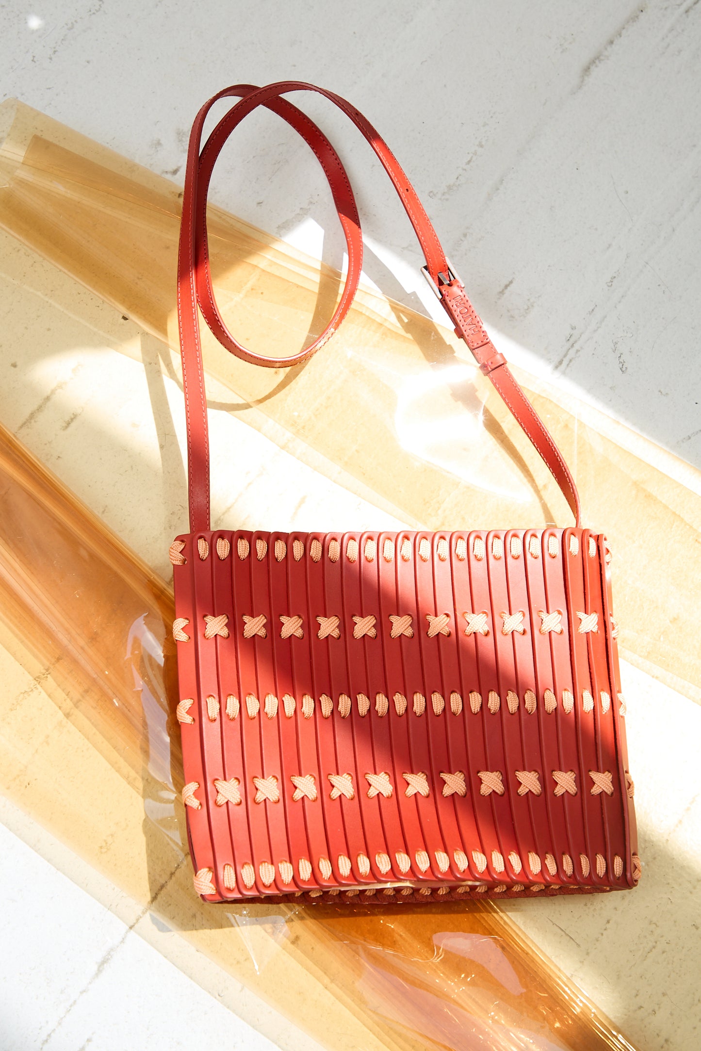 A Hatori Leather Crossbody in Red with Light Coral Lace is placed on a light surface illuminated by sunlight.