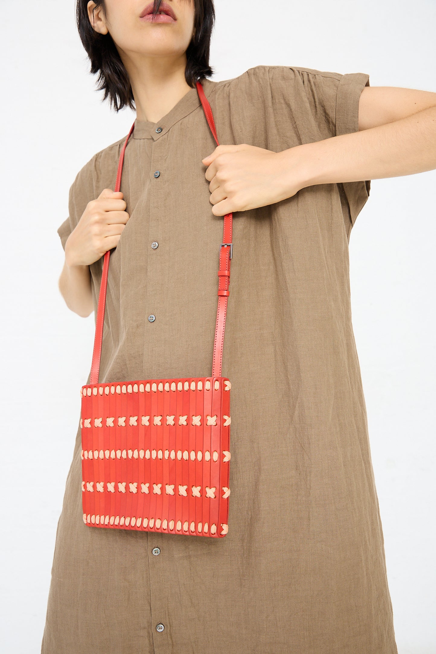 A person holding a Hatori Leather Crossbody in Red with Light Coral Lace over their shoulder. They are wearing a button-up beige dress.