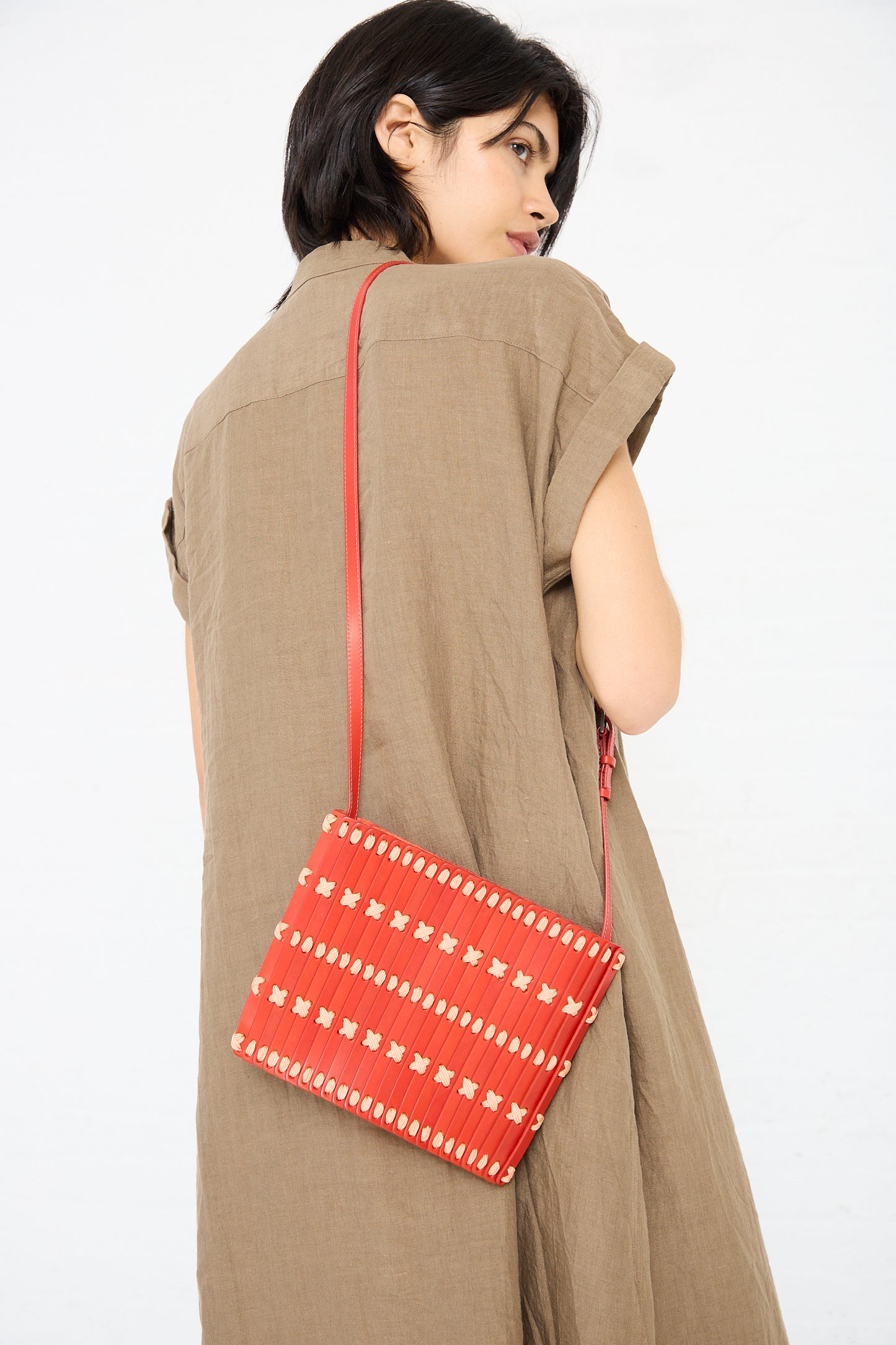 The woman, dressed in a brown dress, is turned slightly away, showcasing the Hatori Leather Crossbody in Red with Light Coral Lace with an adjustable shoulder strap hanging over her shoulder.