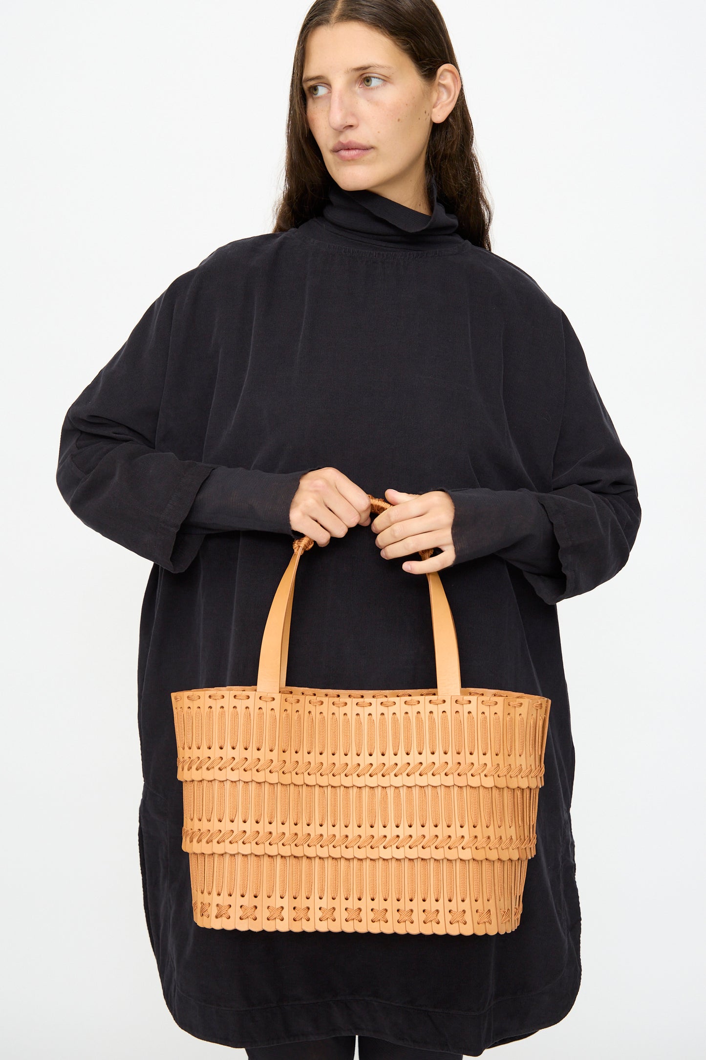 A woman in a loose black dress holds the Hatori Leather Tote 198 in Natural with Natural Lace, featuring two handles and handcrafted from vegetable tanned leather, against a plain white background.