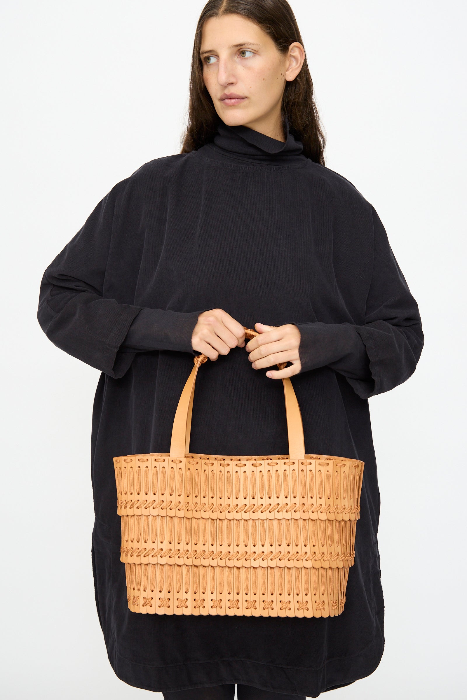 A woman in a loose black dress holds the Hatori Leather Tote 198 in Natural with Natural Lace, featuring two handles and handcrafted from vegetable tanned leather, against a plain white background.