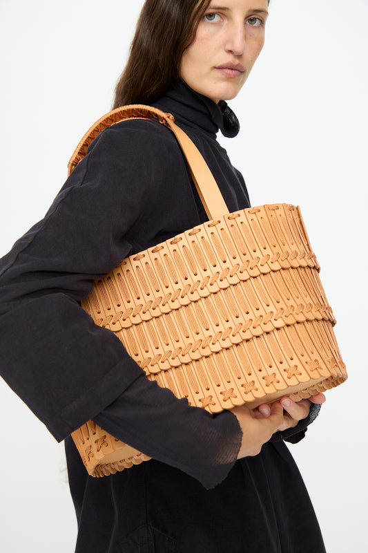 A woman in a black turtleneck carries the Hatori Leather Tote 198 in Natural with Natural Lace on her shoulder, highlighting its handcrafted tan woven detail against a plain white background.