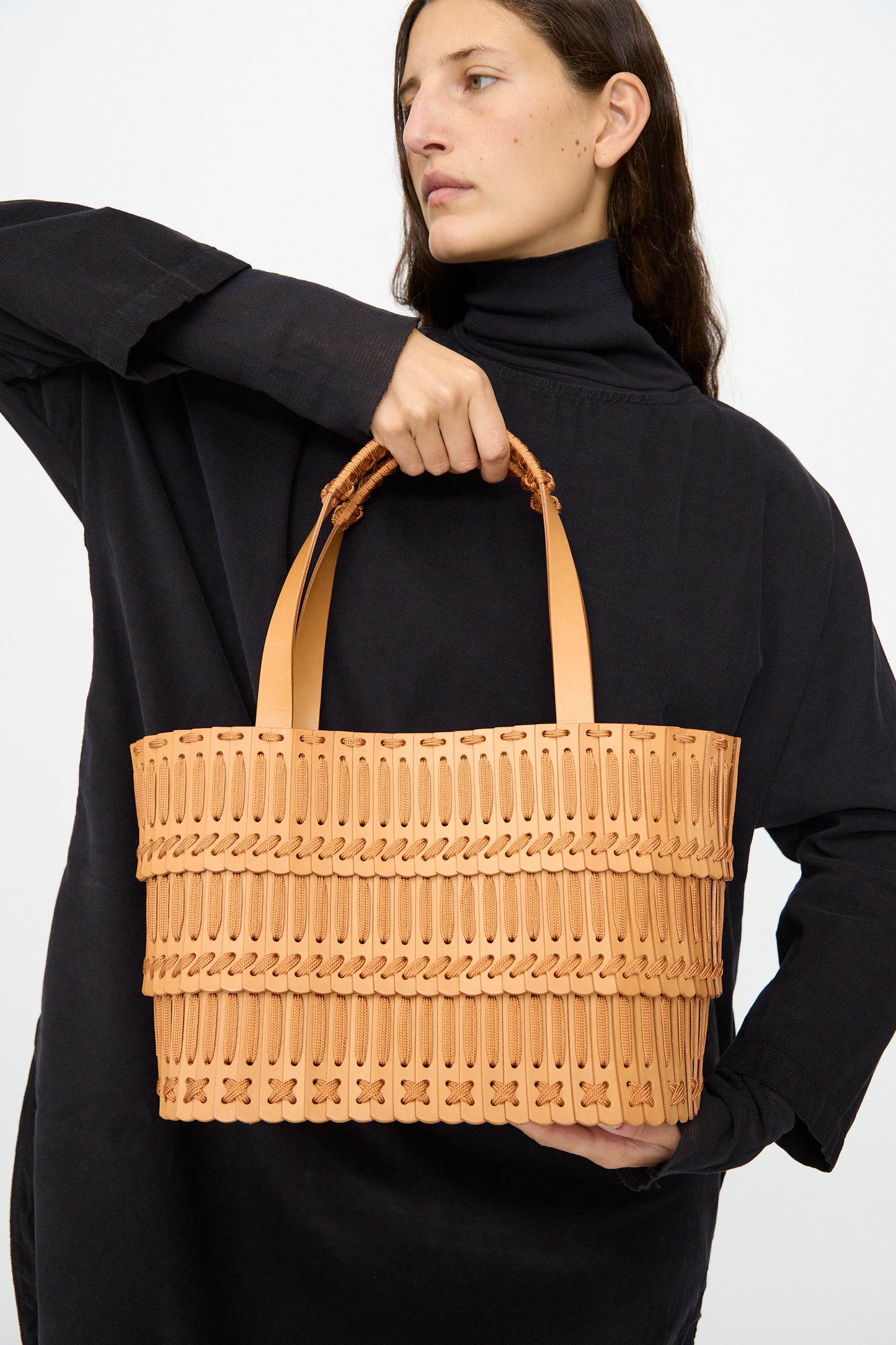 A person in a black long-sleeve top holds the Hatori Leather Tote 198 in Natural with Natural Lace, a large handcrafted tote featuring tan woven leather and two handles, set against a plain background.