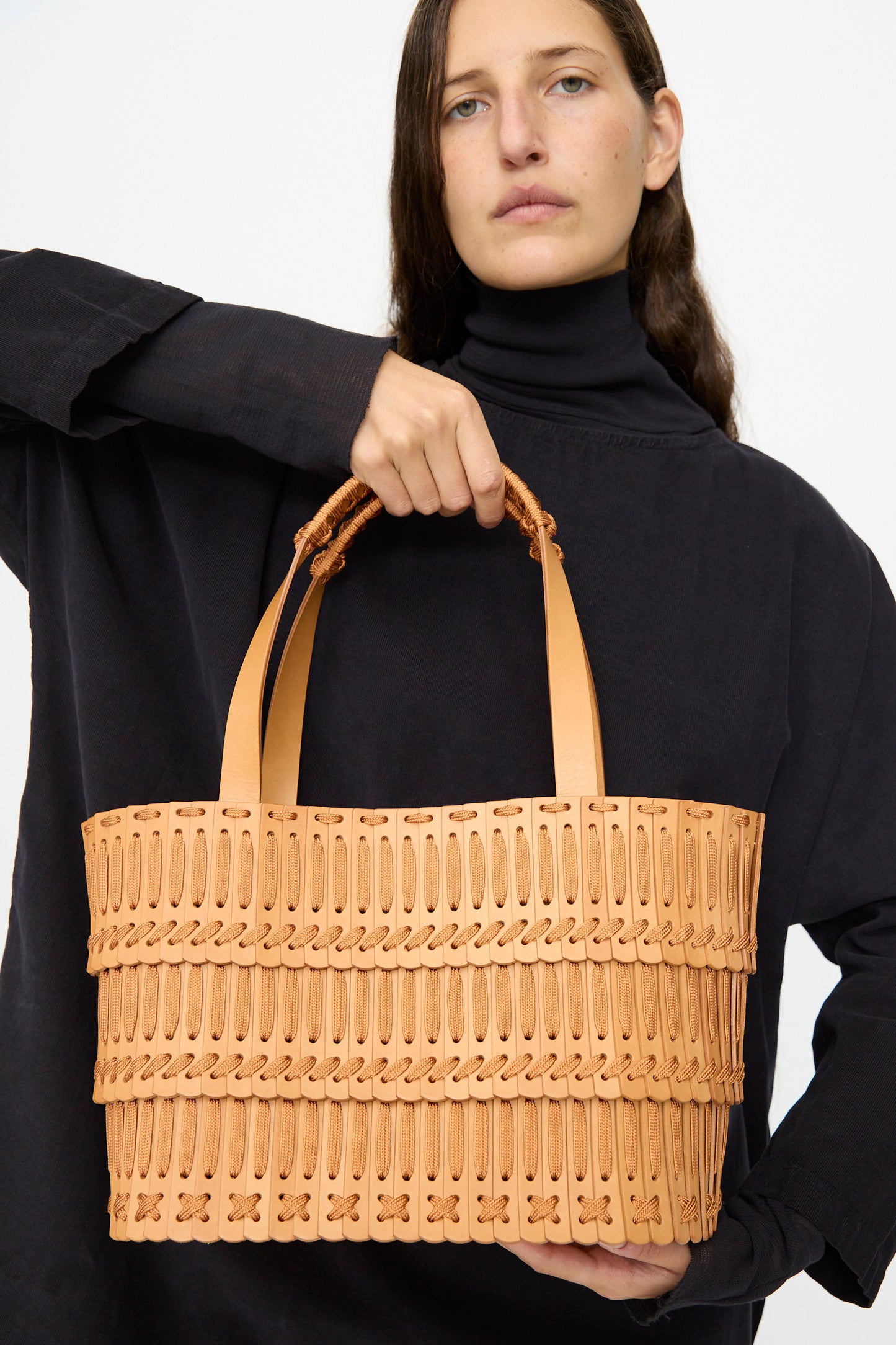 A woman in a black top holds the Hatori Leather Tote 198 in Natural with Natural Lace, featuring two handles and a patterned texture, posing against a plain light background.