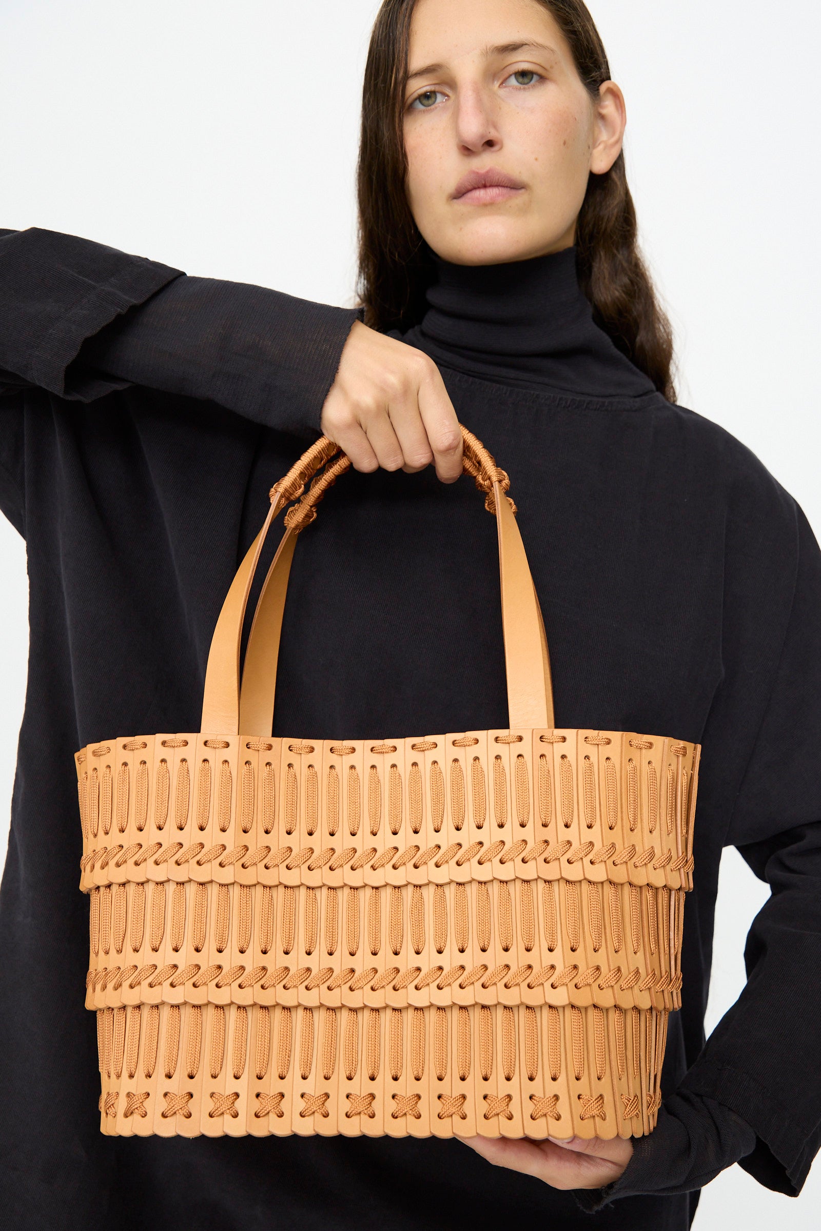 A woman in a black top holds the Hatori Leather Tote 198 in Natural with Natural Lace, featuring two handles and a patterned texture, posing against a plain light background.
