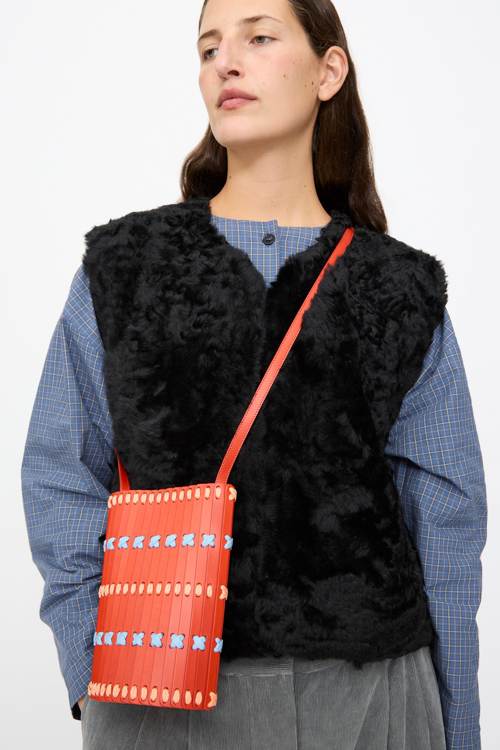 A person wearing a black faux fur vest over a blue plaid shirt, carrying the Hatori Leather Crossbody 30 Artist Edition with Alex Dodge in Red.