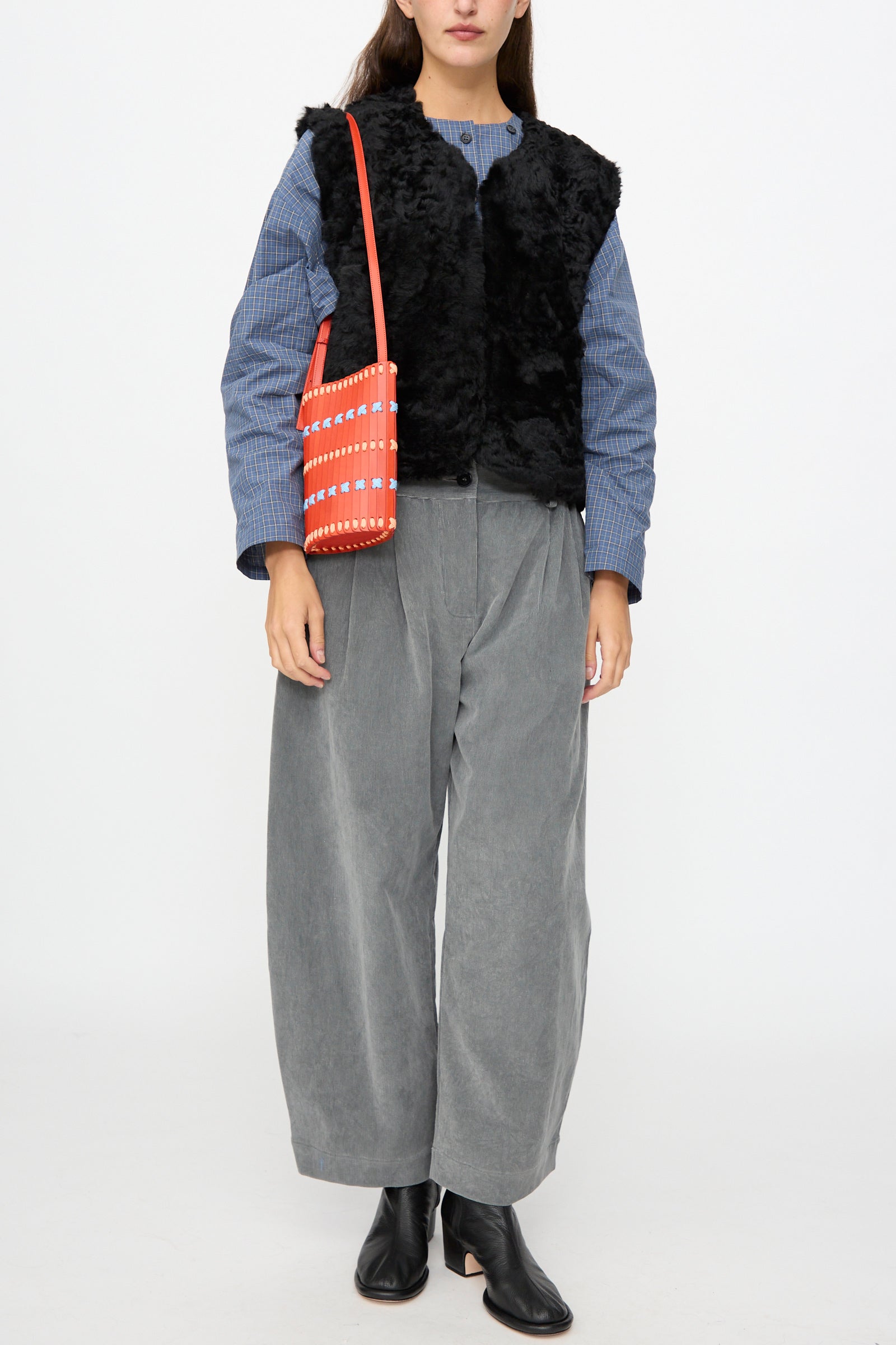 A person wearing a black fur vest, blue-gray striped shirt, gray wide-leg pants, and black boots stands against a white background, carrying the Hatori Leather Crossbody 30 Artist Edition with Alex Dodge in Red—handcrafted in Japan from vegetable-tanned leather.