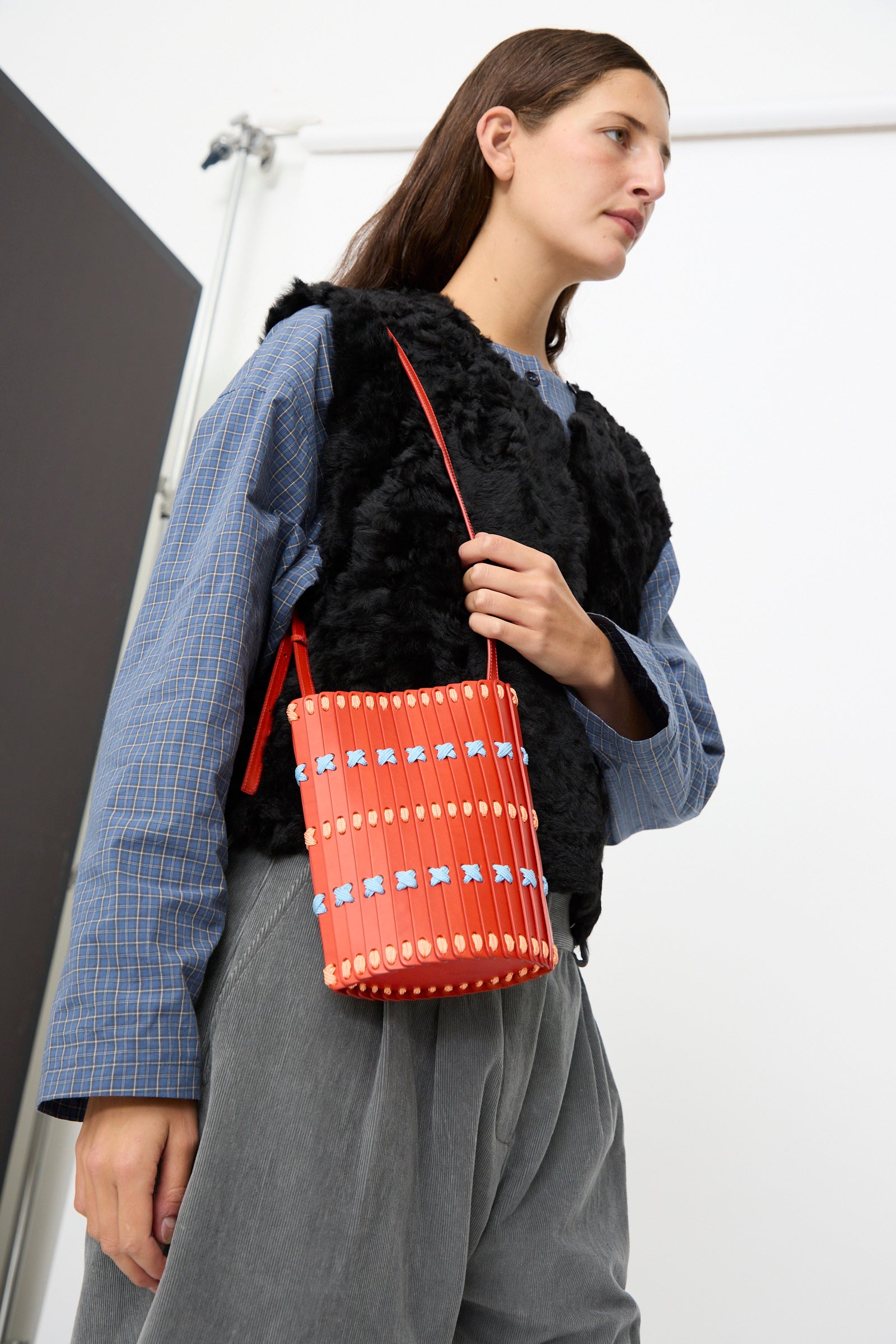 A person wearing a blue plaid shirt, black furry vest, and gray pants holds the Hatori Leather Crossbody 30 Artist Edition with Alex Dodge in Red—a vibrant bucket-style shoulder bag handcrafted in Japan from vegetable tanned leather, featuring bold red, blue, and white accents.