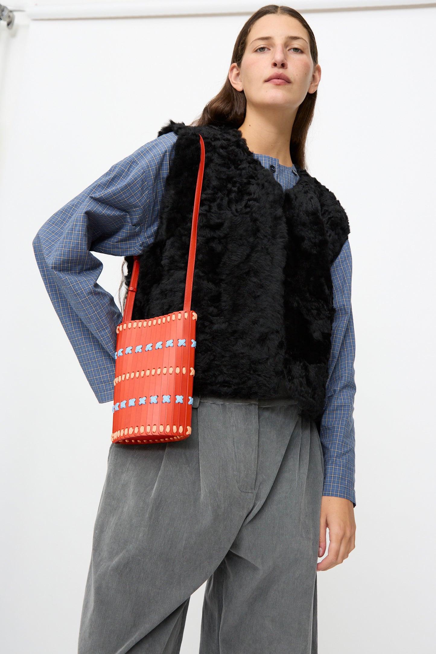 A person wearing a blue plaid shirt, black faux fur vest, gray wide-leg pants, and carrying the Hatori Leather Crossbody 30 Artist Edition with Alex Dodge in Red stands against a plain white background.