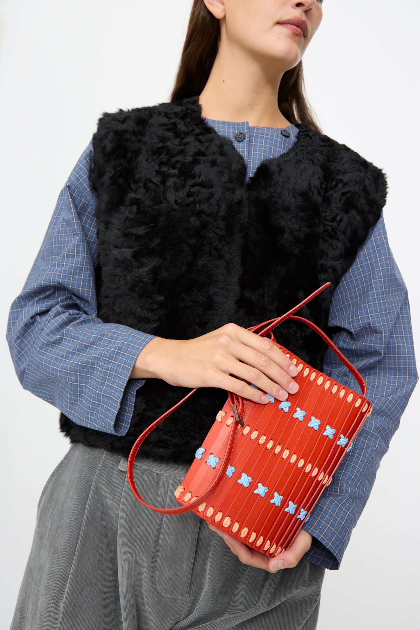 Woman wearing a black faux fur vest, blue checkered shirt, and gray pants, holding the Hatori Leather Crossbody 30 Artist Edition with Alex Dodge in Red.