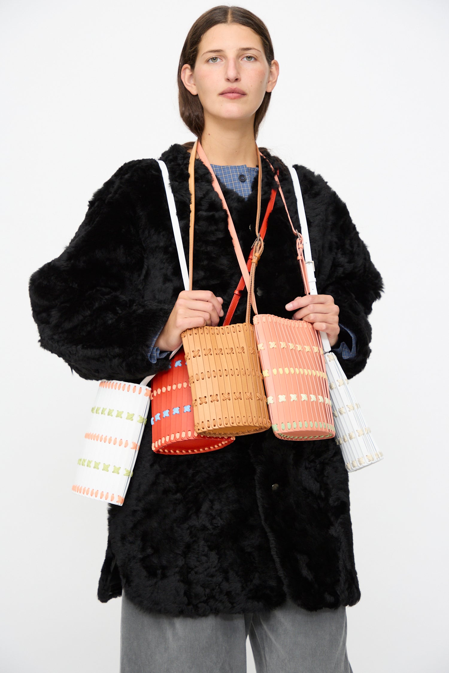 A person dressed in a black faux fur coat is holding and wearing several vibrant woven handbags, including the Hatori Leather Crossbody 30 in Pink with Multi Lacing—a handcrafted shoulder bag made in Japan—draped around their neck and hands against a plain background.
