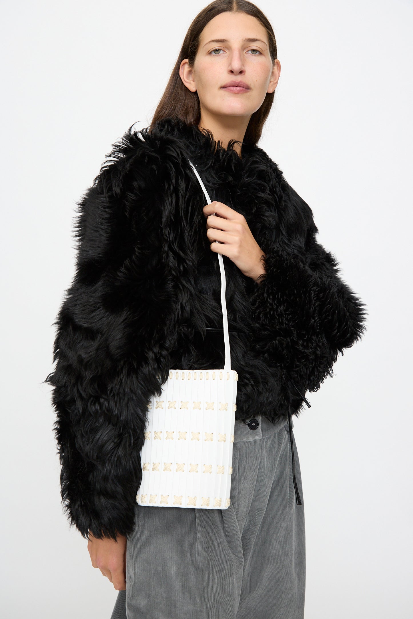 A person wearing a black faux fur jacket and gray pants, holding the Hatori Leather Crossbody 30 in White with White Lacing, stands against a plain white background.